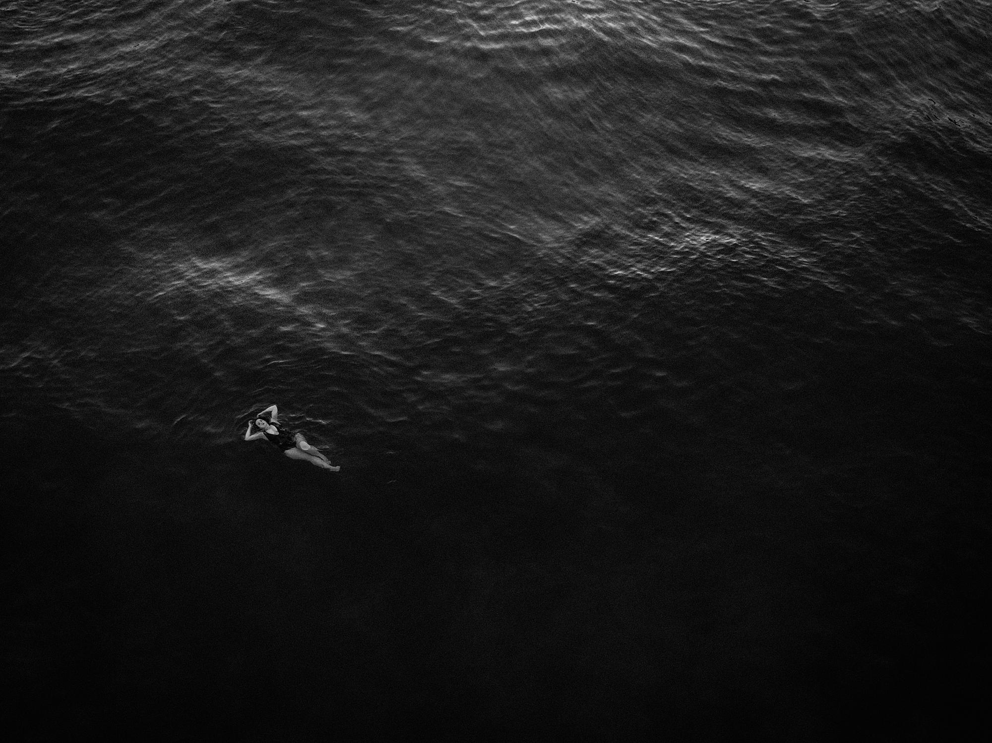 blue hour portrait ideas with drone boudoir portrait in black and white featuring a woman laying on her back floating in the ocean on maui