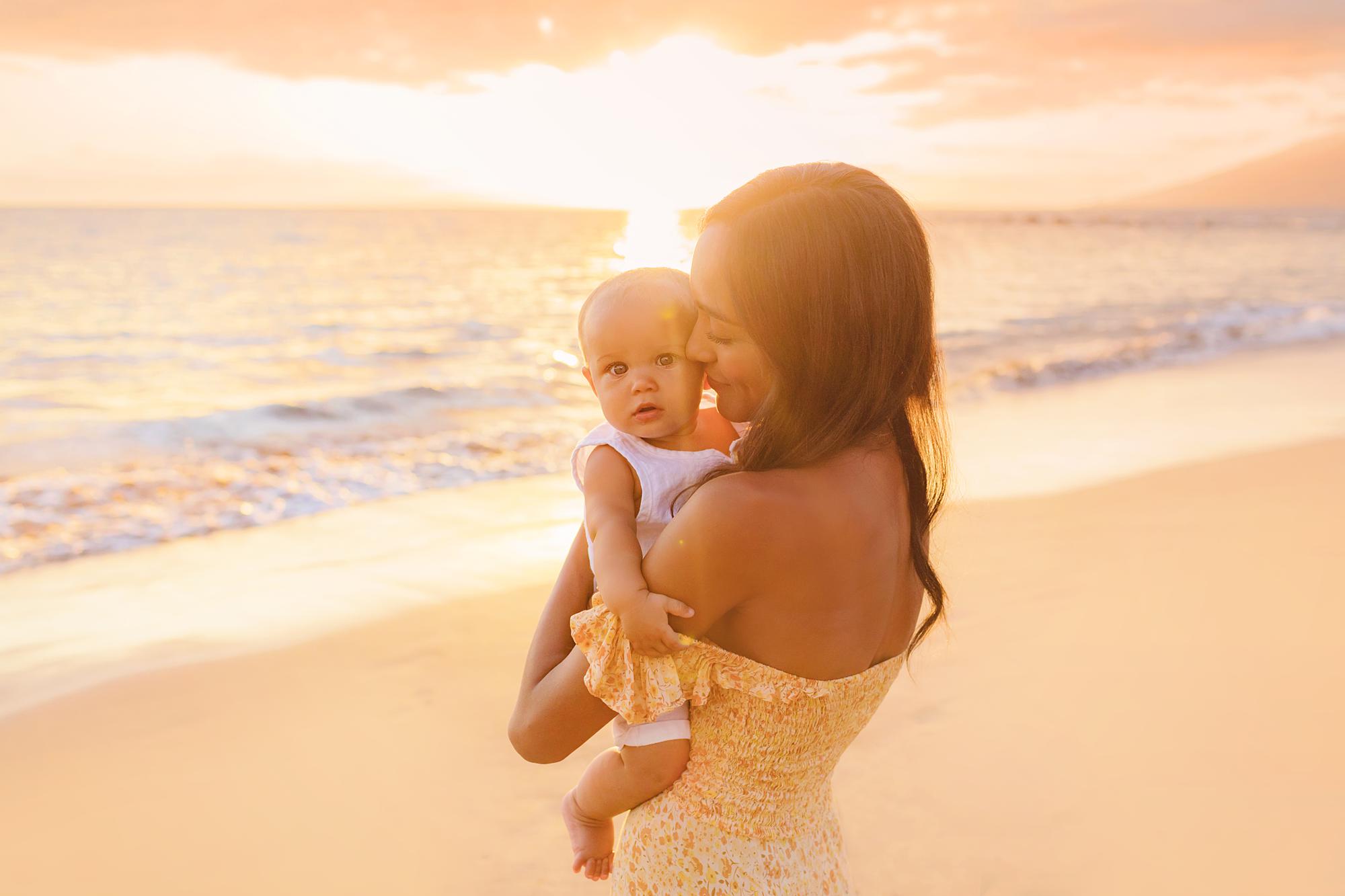Mother kisses her baby's cheek as he looks at the camera while the sun sets during their photoshoot on Maui
