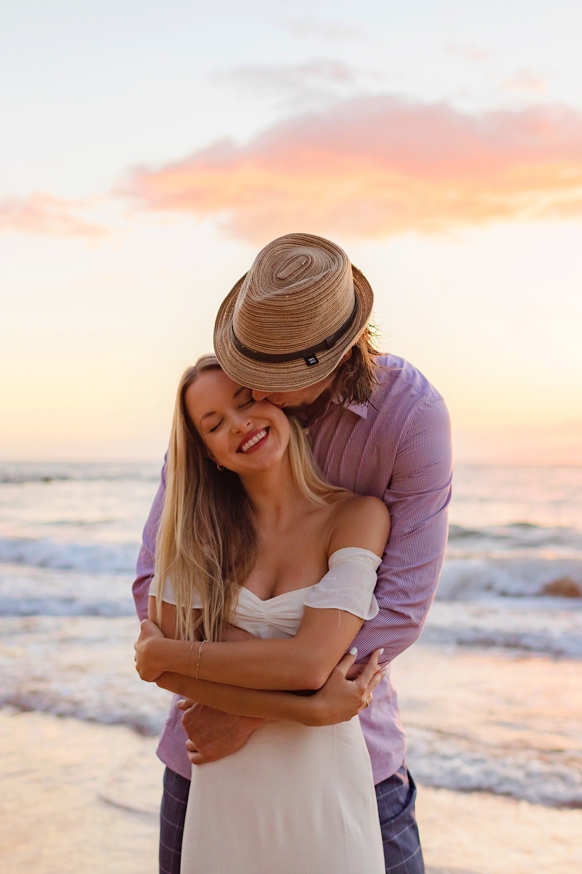 beautiful woman in white dress smiling as fiance in hat embraces her and kisses her cheek at sunset on Maui.