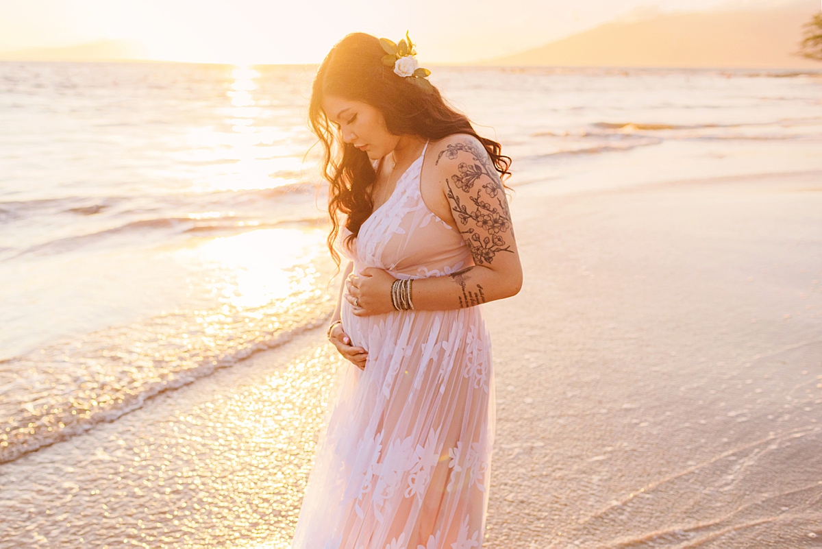 White maternity gown worn by expecting mother looking down at belly at sunset