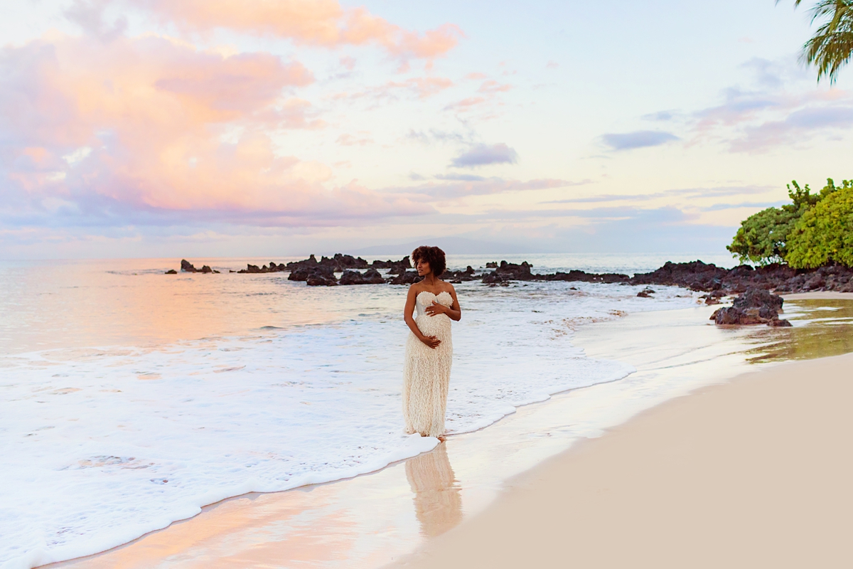 A pregnant woman wearing a cream gown stands in the water on the beach during her babymoon in Maui, Hawaii. 