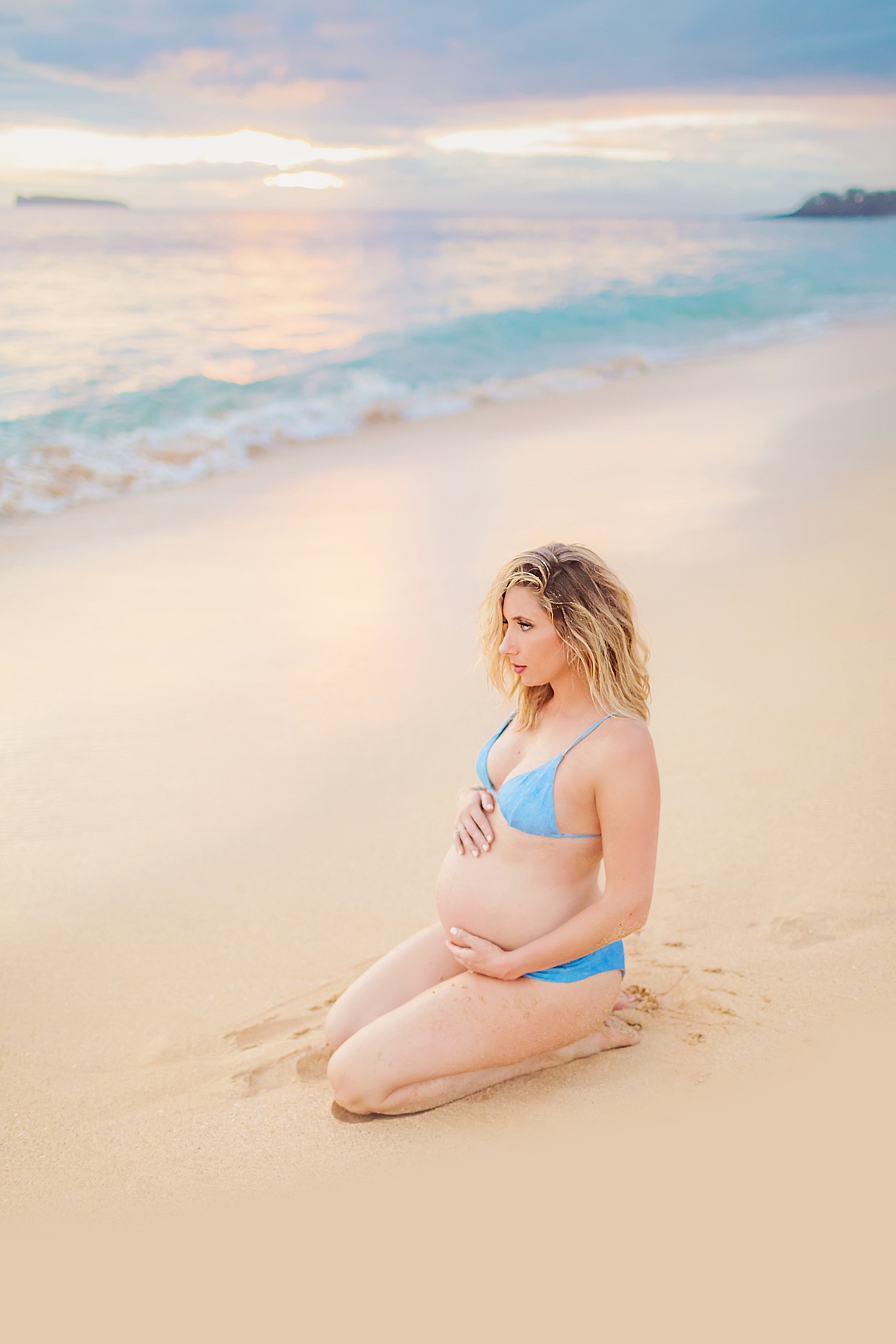 Blond pregnant woman in blue bikini kneeling on the beach