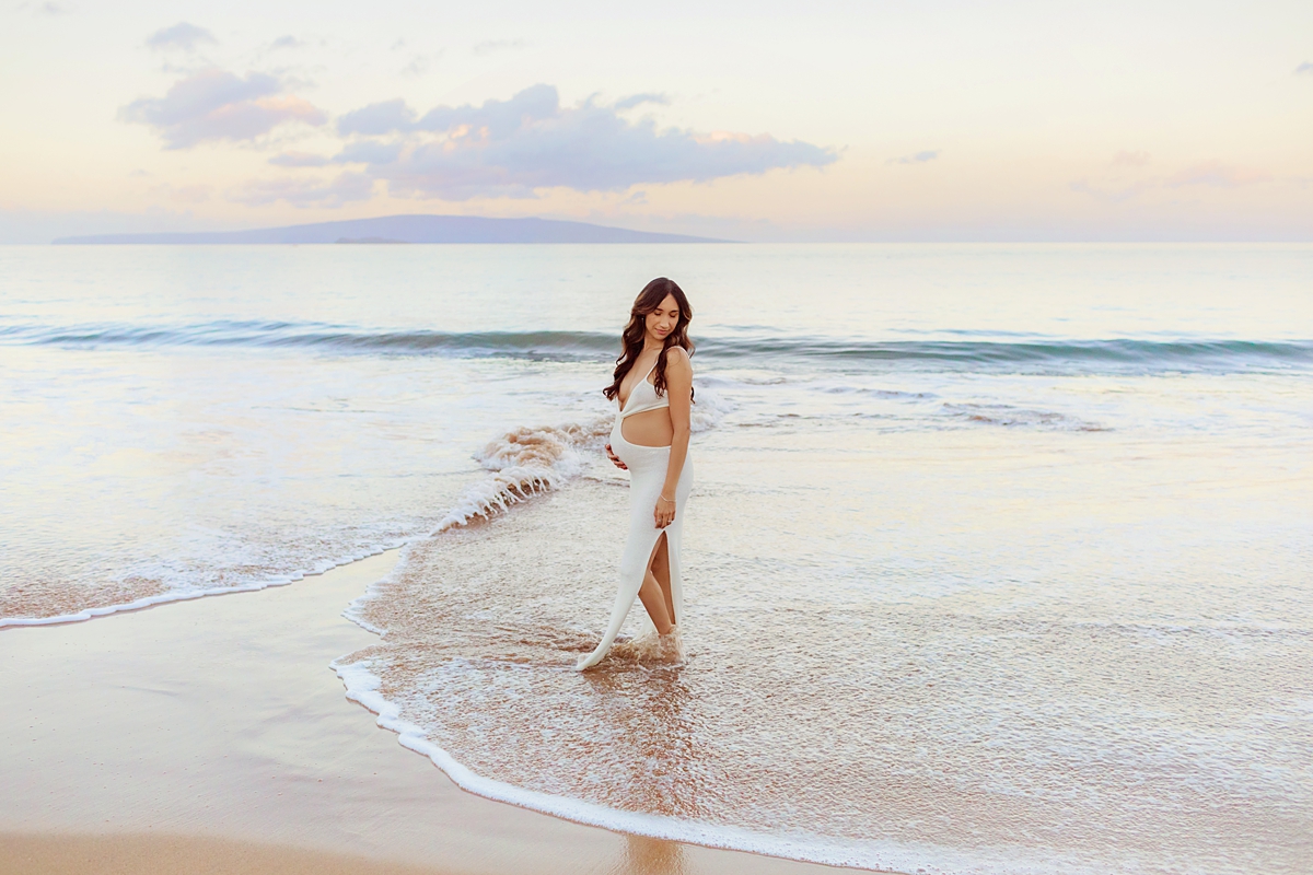 Pregnant woman in long white cut-out dress holds her belly and glances behind her while walking on the beach