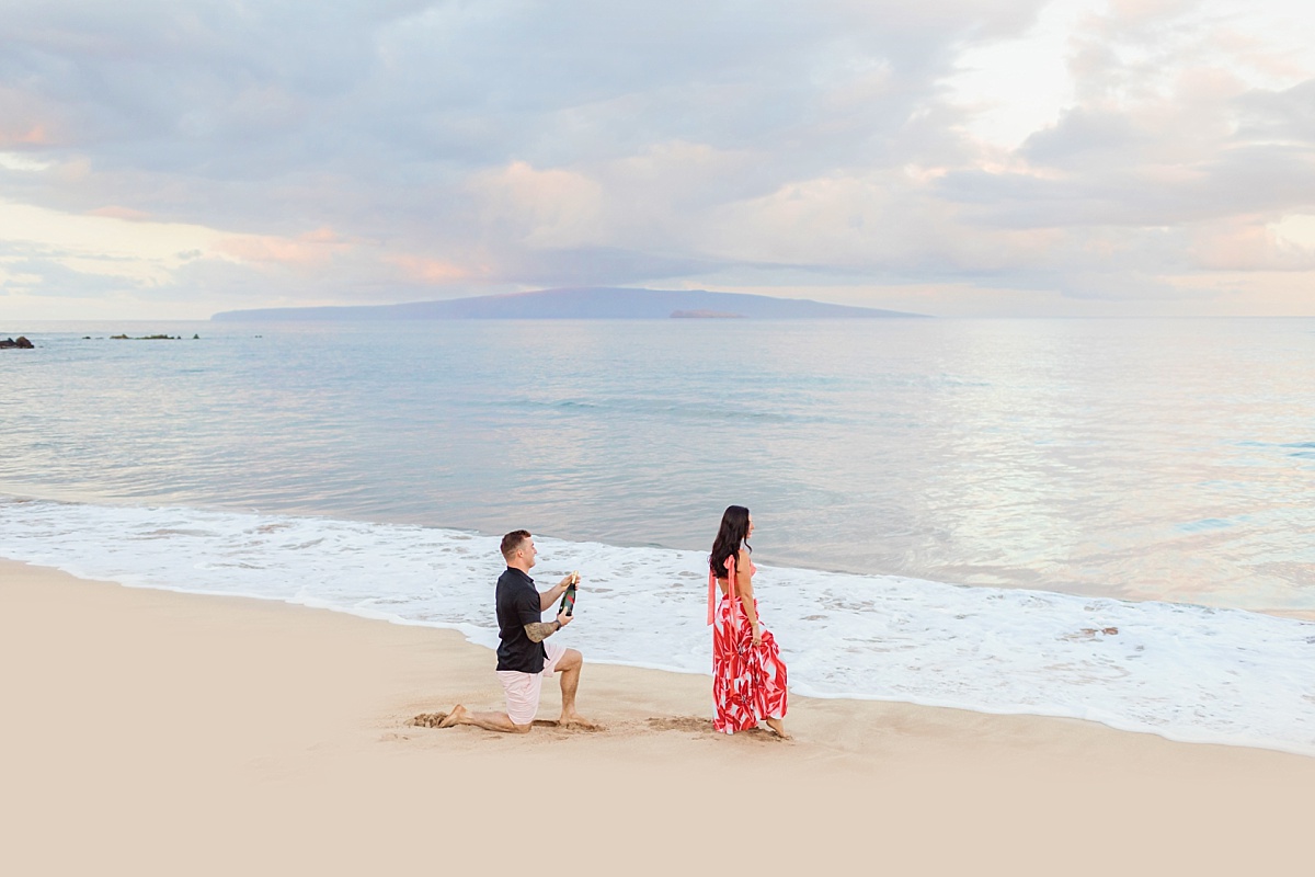 A man holding a bottle of champagne down on one knee while his girlfriend looks out at the ocean, about to propose on the beach in Wailea, Maui.