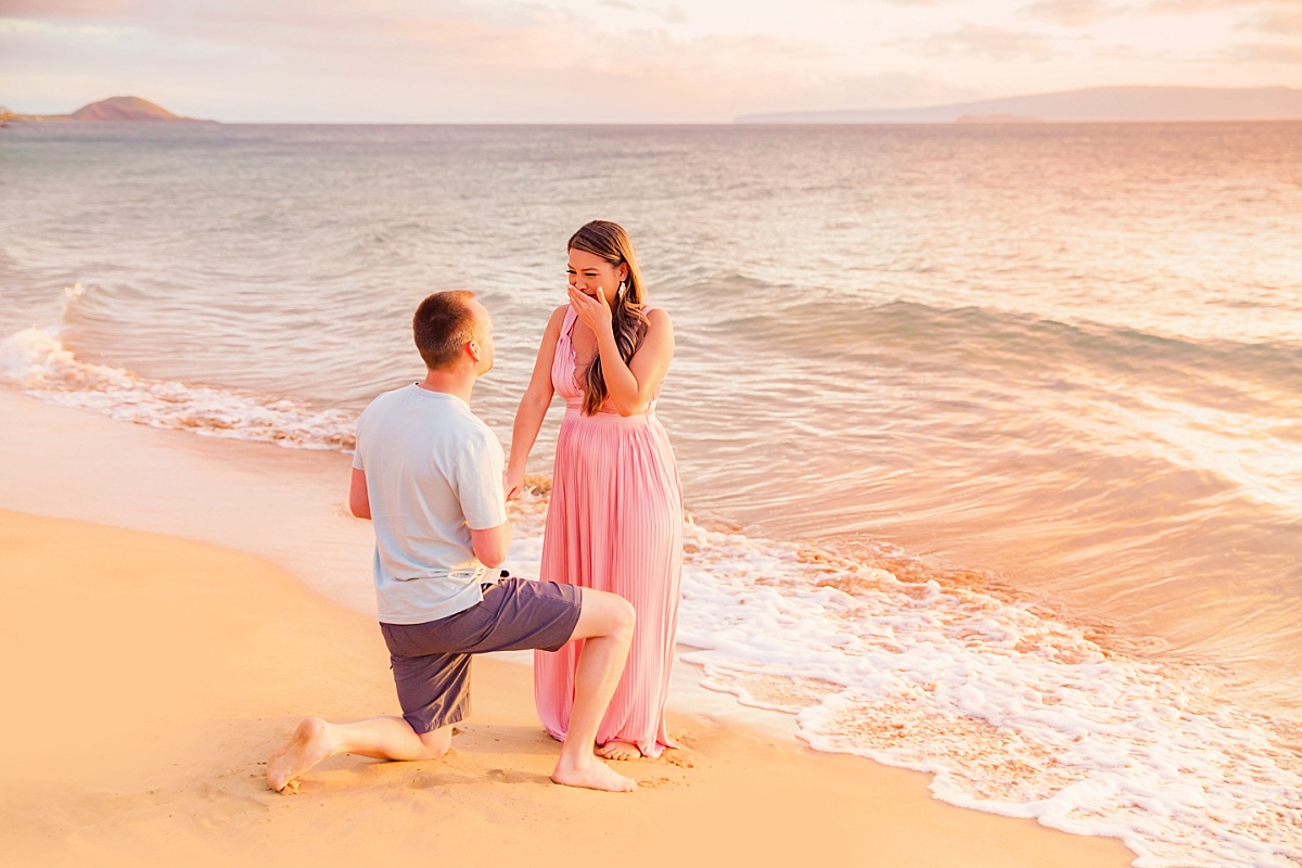 A woman in a flowing pink dress covers her mouth in happy surprise as a man proposes on the beach at sunrise in Hawaii. 