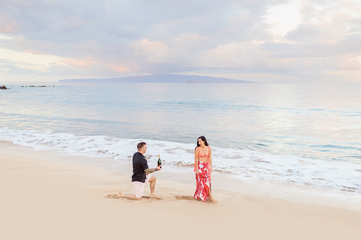 A happily surprised woman turns to see her boyfriend down on one knee with a bottle of champagne during a beach proposal in Hawaii.