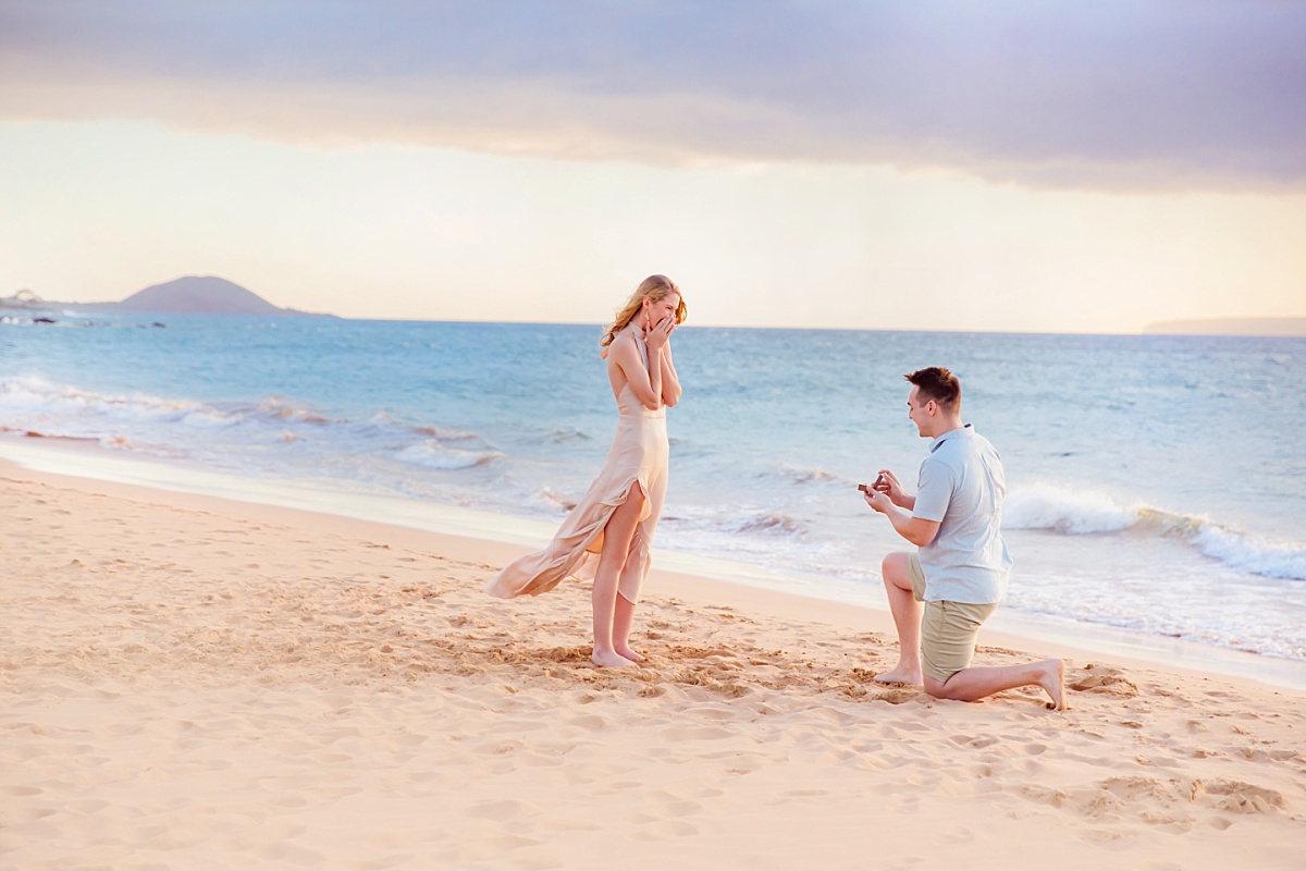 A beautiful woman is surprised while a man presents a ring down on one knee during a beach proposal in Maui, and the moment is captured by Wailea engagement photographers Love + Water photography.