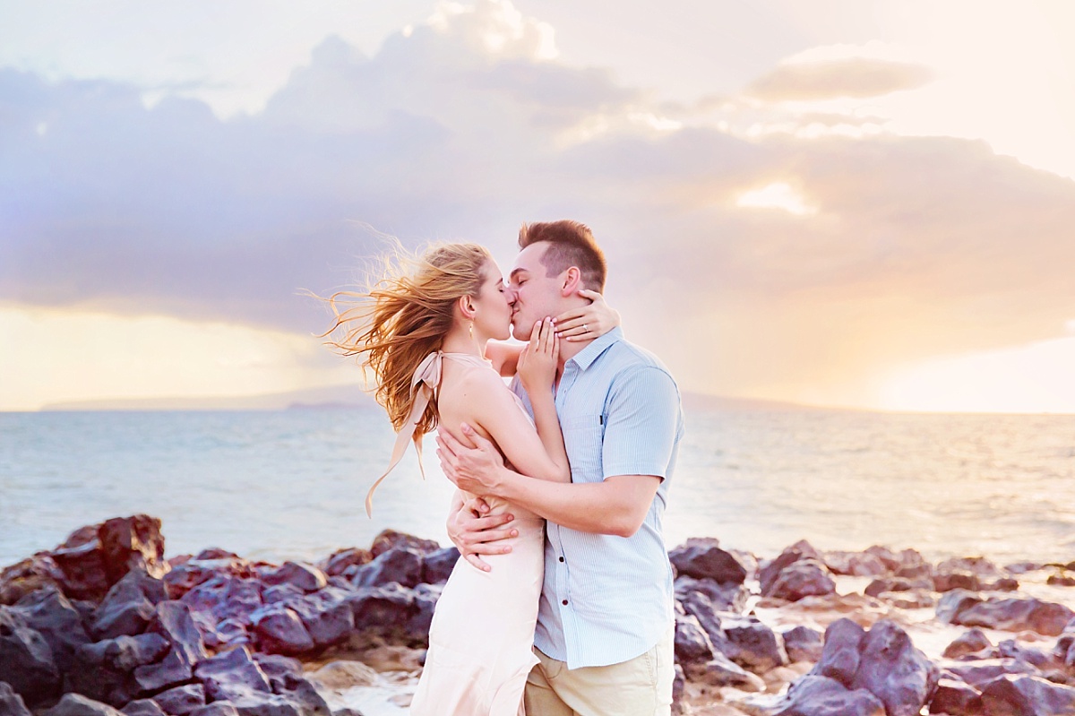 A beautiful couple holds each other and kisses in front of the ocean during a beach engagement session in Hawaii.