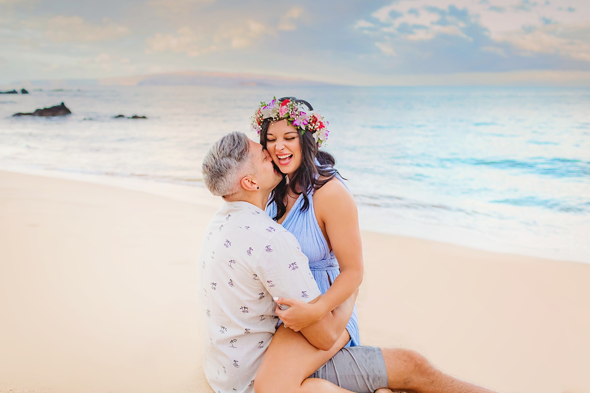 Woman in blue gown sits on husbands lap in the sand and laughs as he kisses her cheek