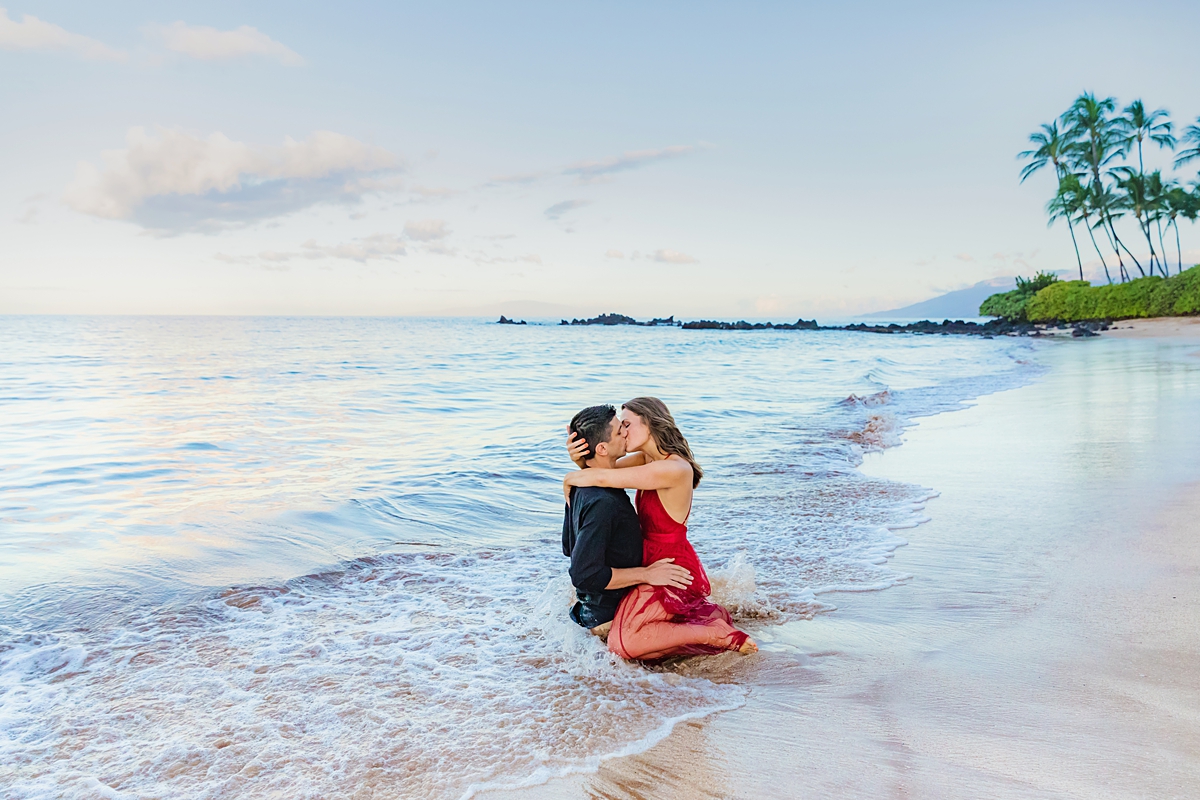 Couple making out in the sand during their beach engagement shoot with palmtrees in the background.