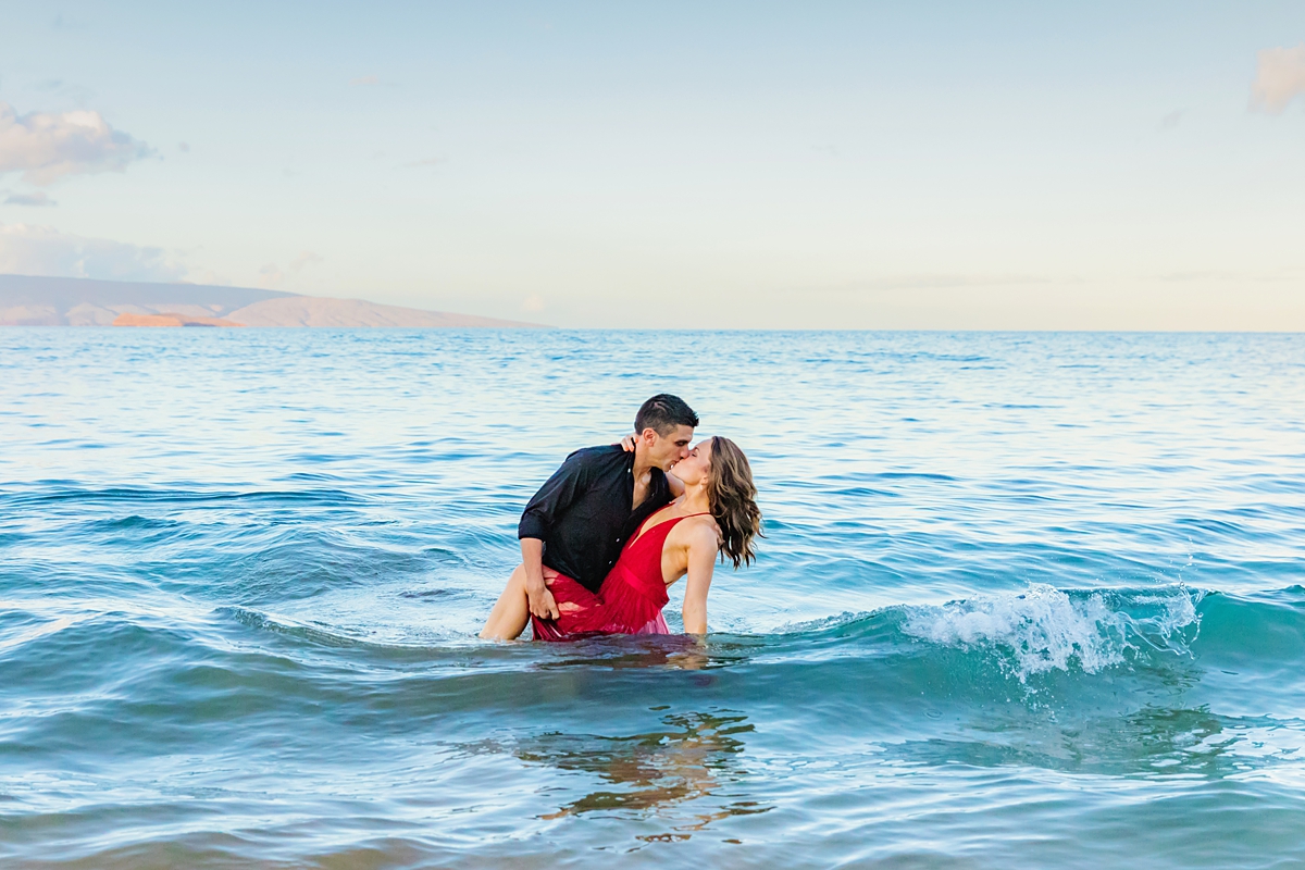 Man dipping his wife in the ocean and kissing her. Woman wears a bright red dress at sunrise.
