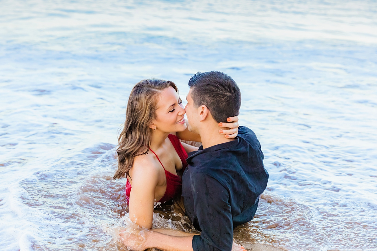 Couple laying on the sand as wave washes up on them before they kiss during a Maui proposal photography session in Wailea.