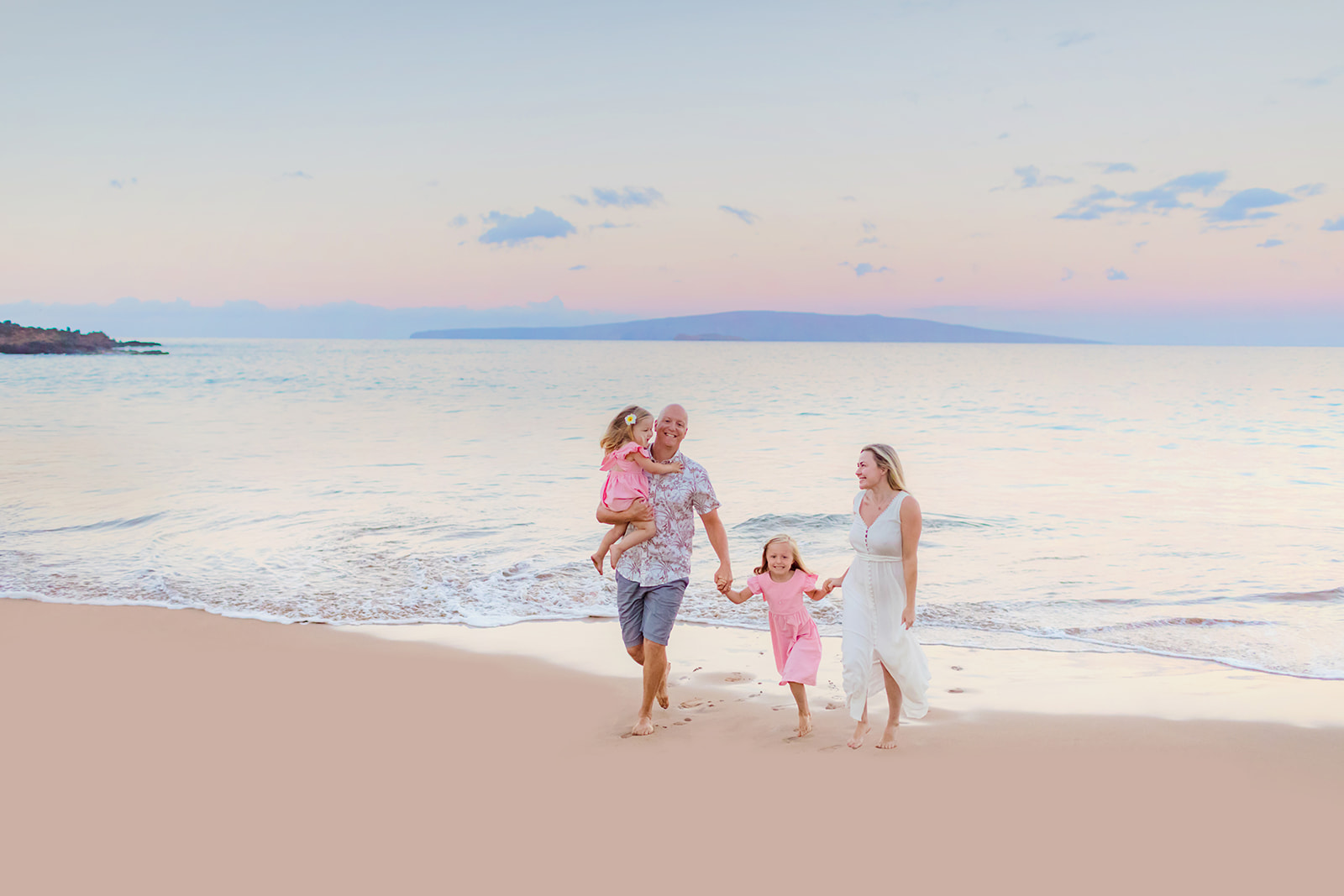 Family of four run toward the camera on the beach at sunrise with still ocean and pink and purple sky
