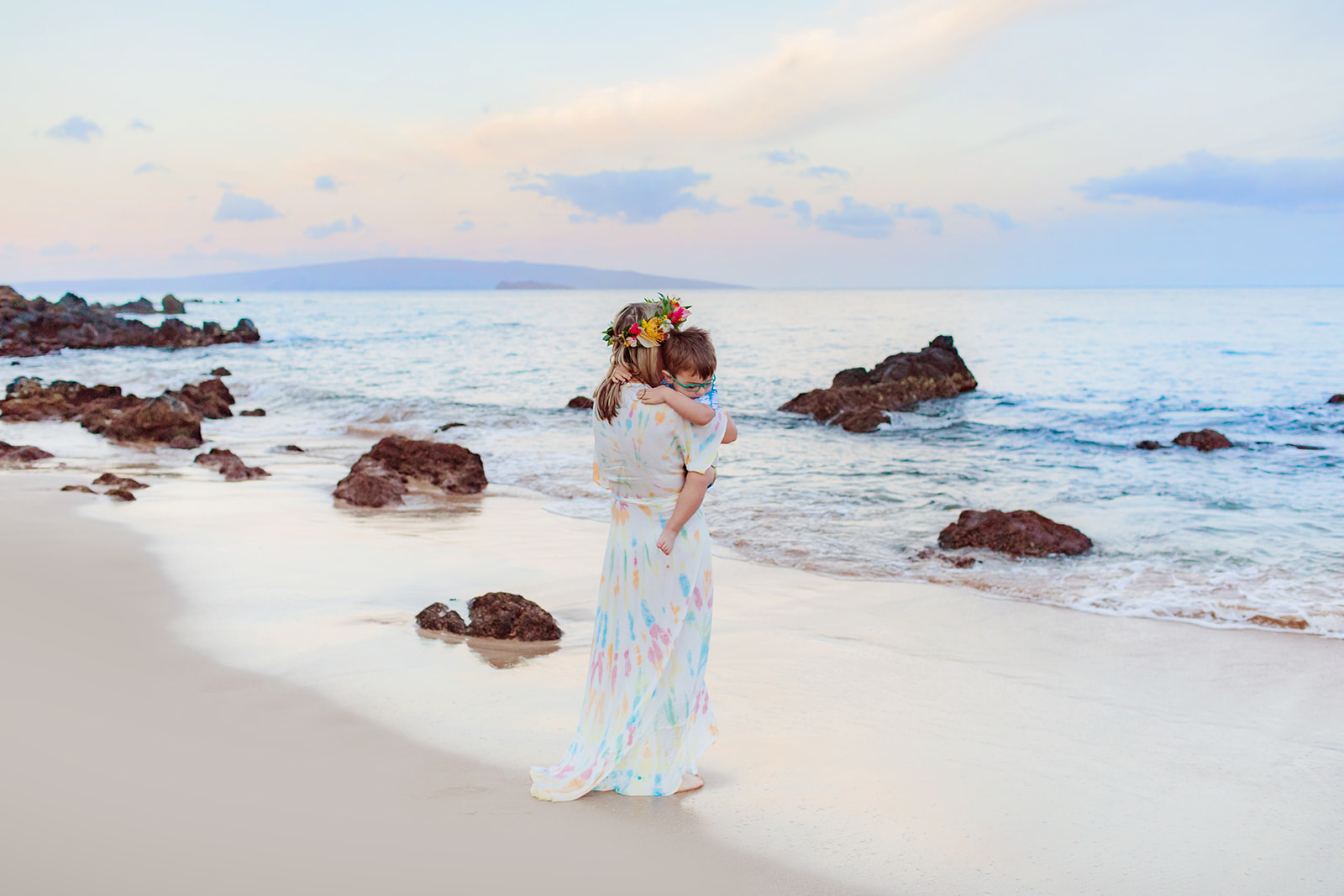 Mother wearing colorful long dress and lei po'o holds son while walking toward the ocean in Hawaii