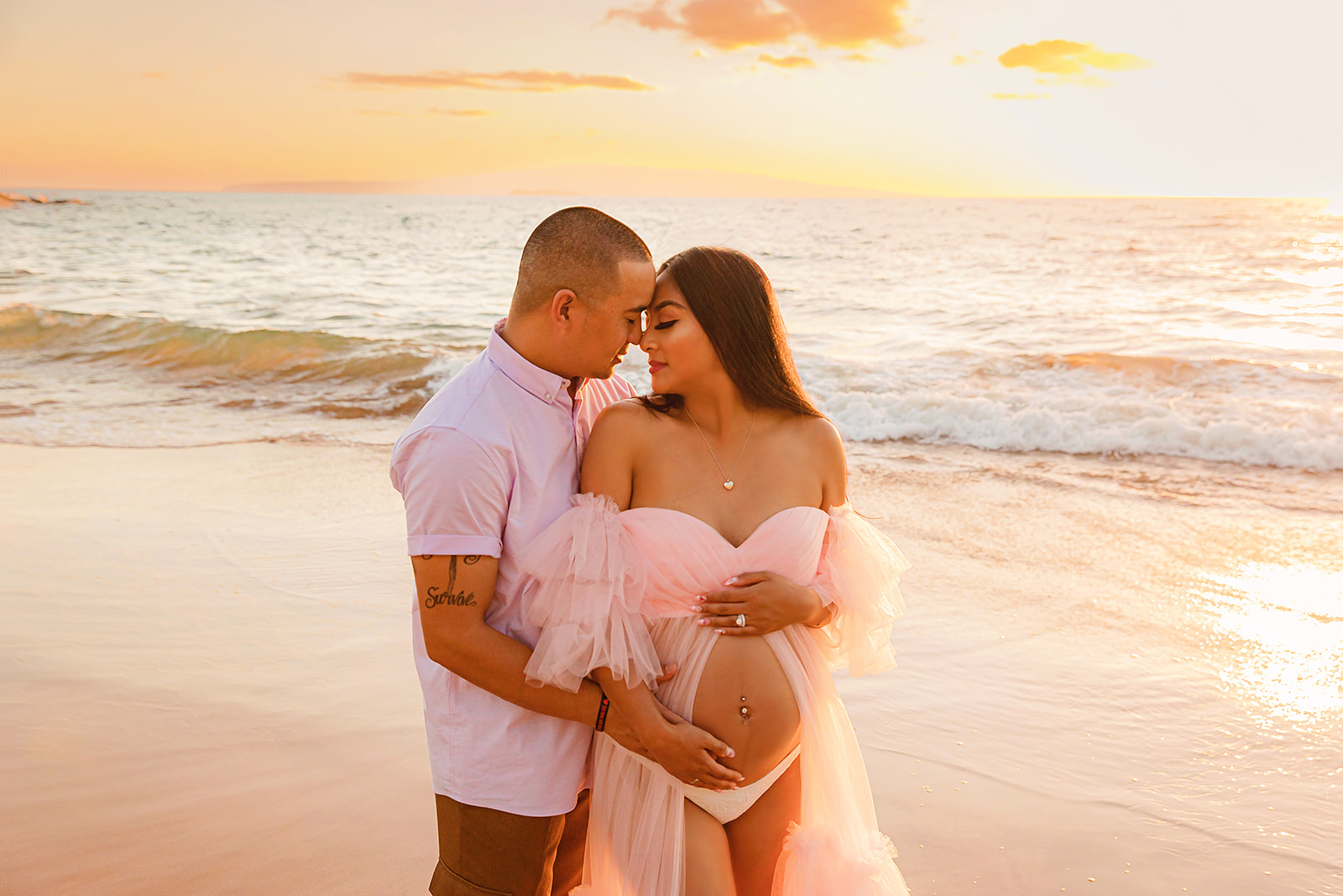 A pregnant woman in a beautiful pink maternity dress stands with her husband for a beach maternity photo session, of one best babymoon ideas in Maui.