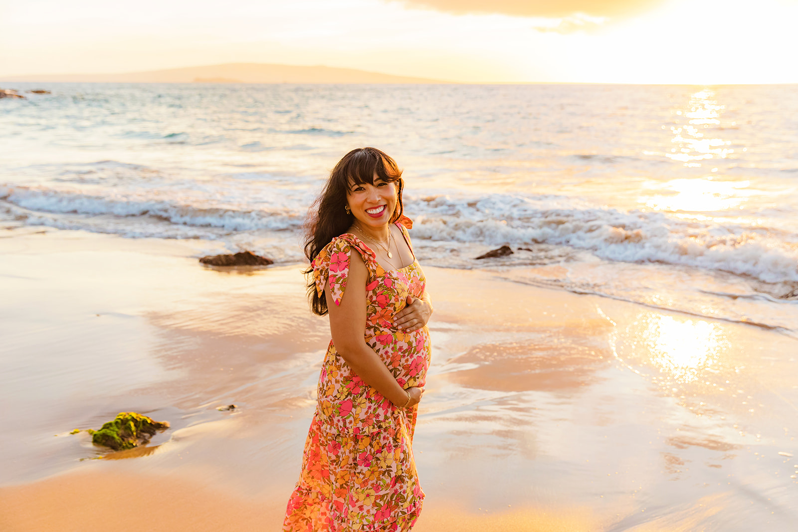 A pregnant woman in a floral dress holds her belly and smiles for maternity photos with Love + Water Photography during a babymoon in Wailea, Maui. 