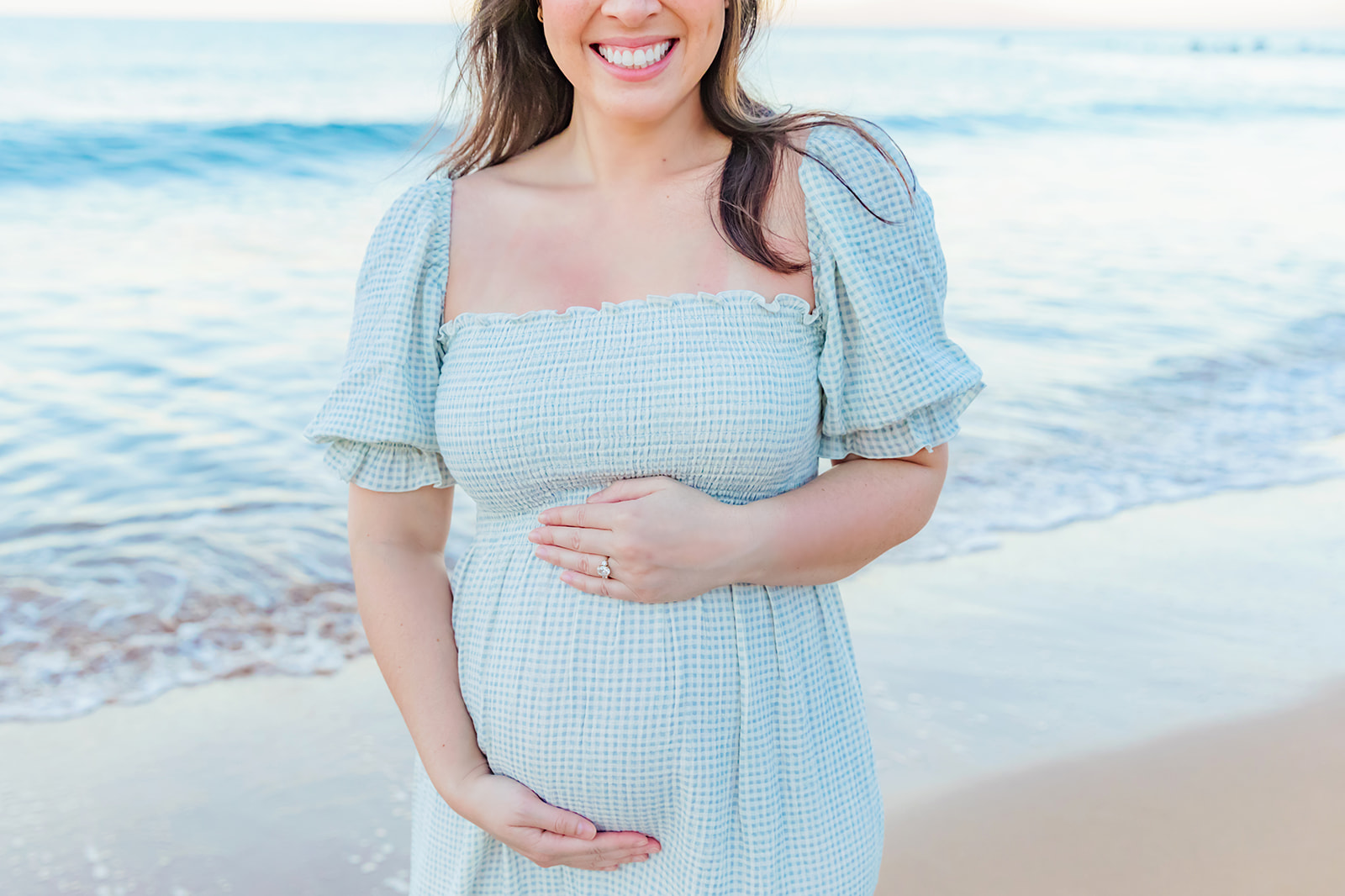 A pregnant woman smiles while holding her belly during a babymoon photo session with Love + Water Photography in Wailea, Maui. 