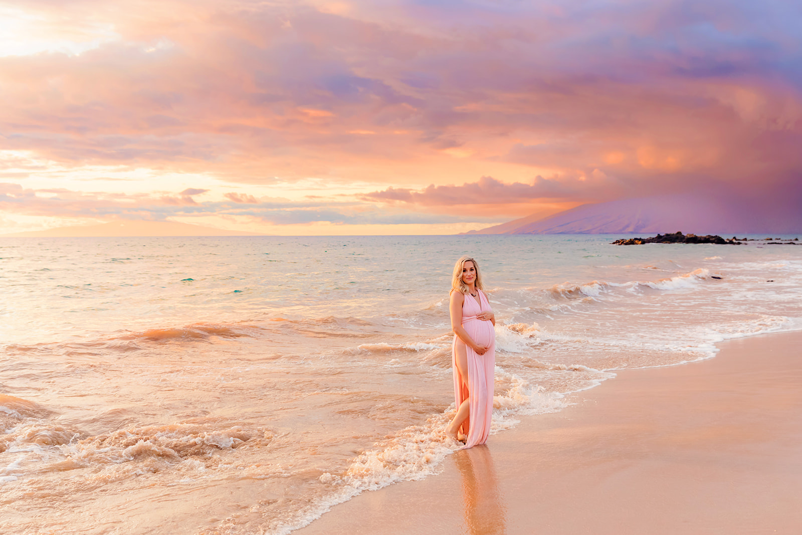 A woman in a pink dress holds her belly while posing for beach maternity photos at sunset during a babymoon in Hawaii.