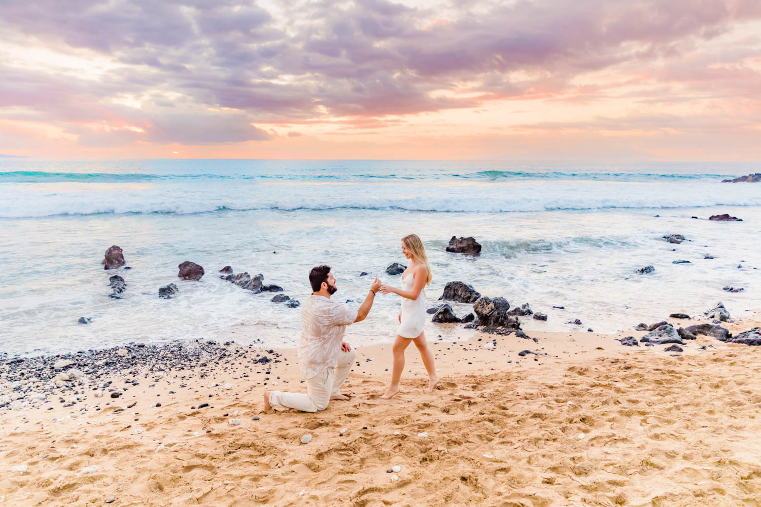 A man gets down on one knee in front of his partner for a beach proposal on Maui, while the moment is captured by Love + Water Photography.