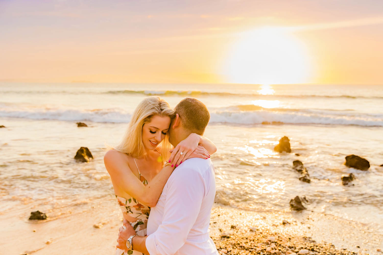 A man and woman embrace as the sun sets on the ocean behind them during a couples beach photoshoot on Maui.