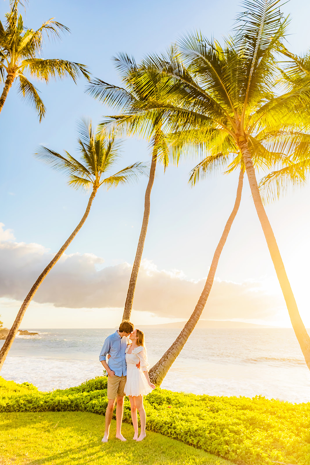 Mia Maples wearing a white dress kisses her fiance wearing a blue button-up shirt and khaki shorts during their engagement photo session in Maui.