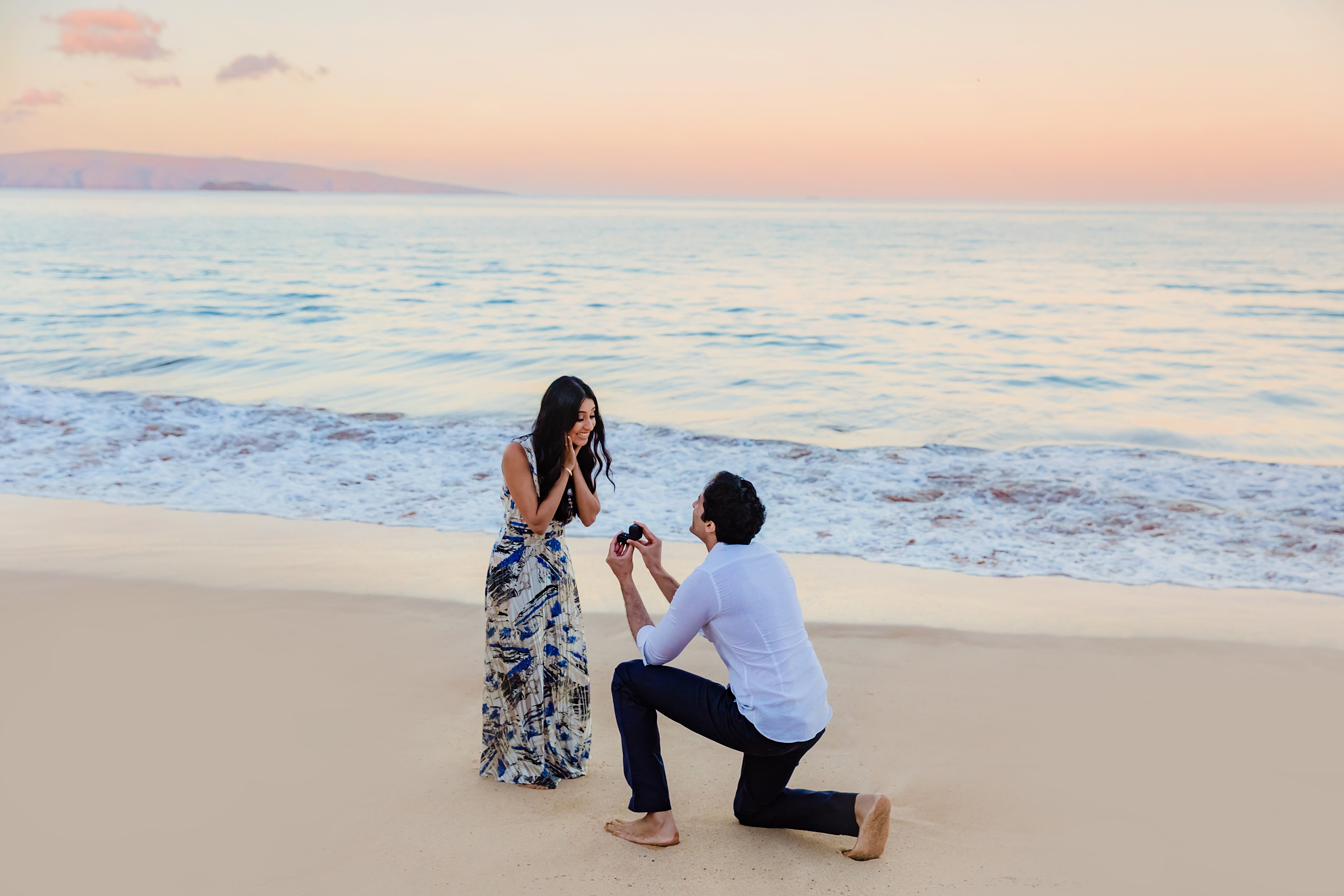 Man in white longsleeve collared shirt and black pants takes a knee to propose to his girlfriend who has long black hair and wears a long patterned dress. Maui surprise proposal photographed by Love and Water Photography.