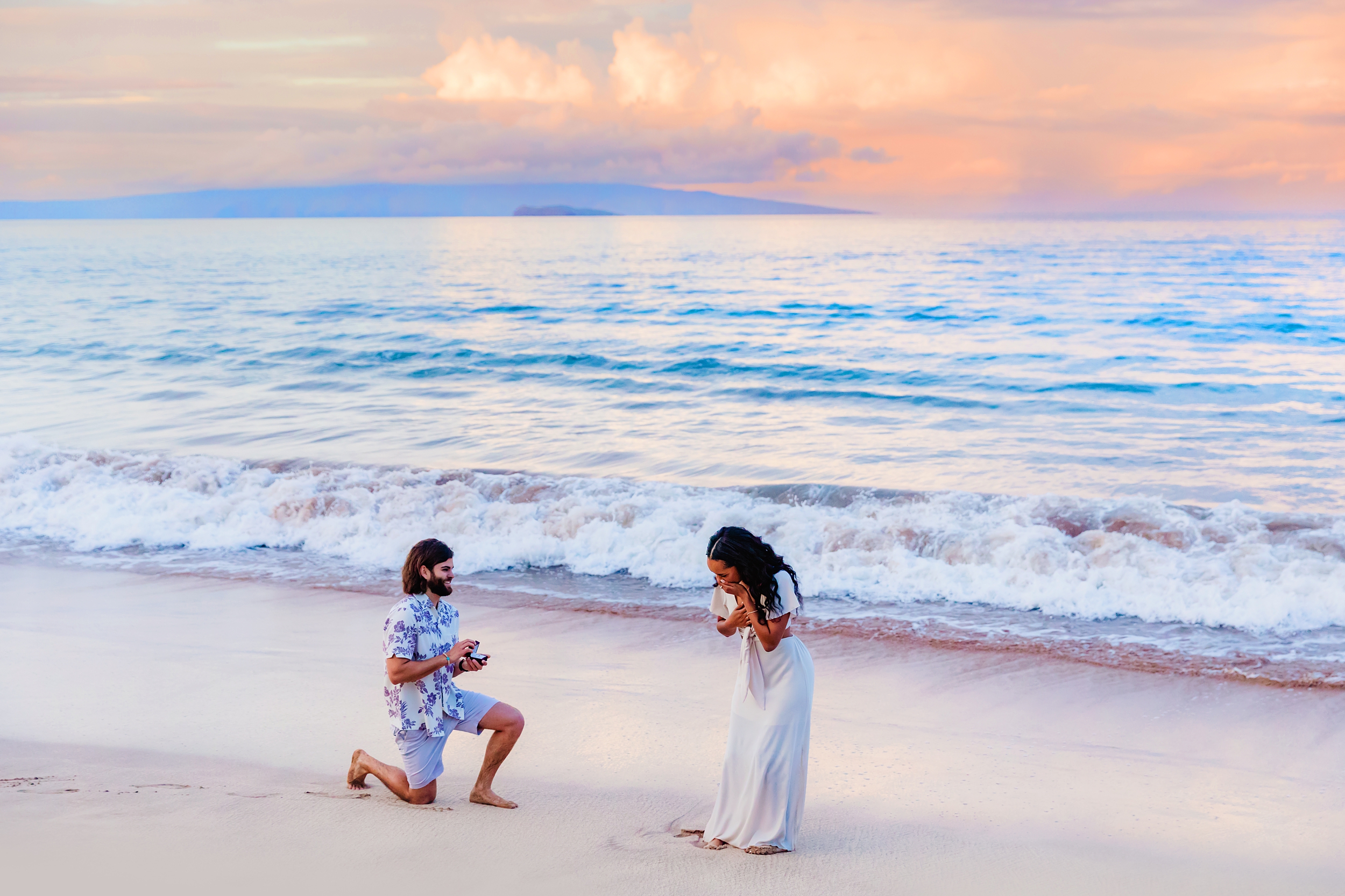 Woman in long white dress holds her mouth and gasps in surprise as her boyfriend proposes to her on the beach in Maui. Photographed at sunrise in Wailea.