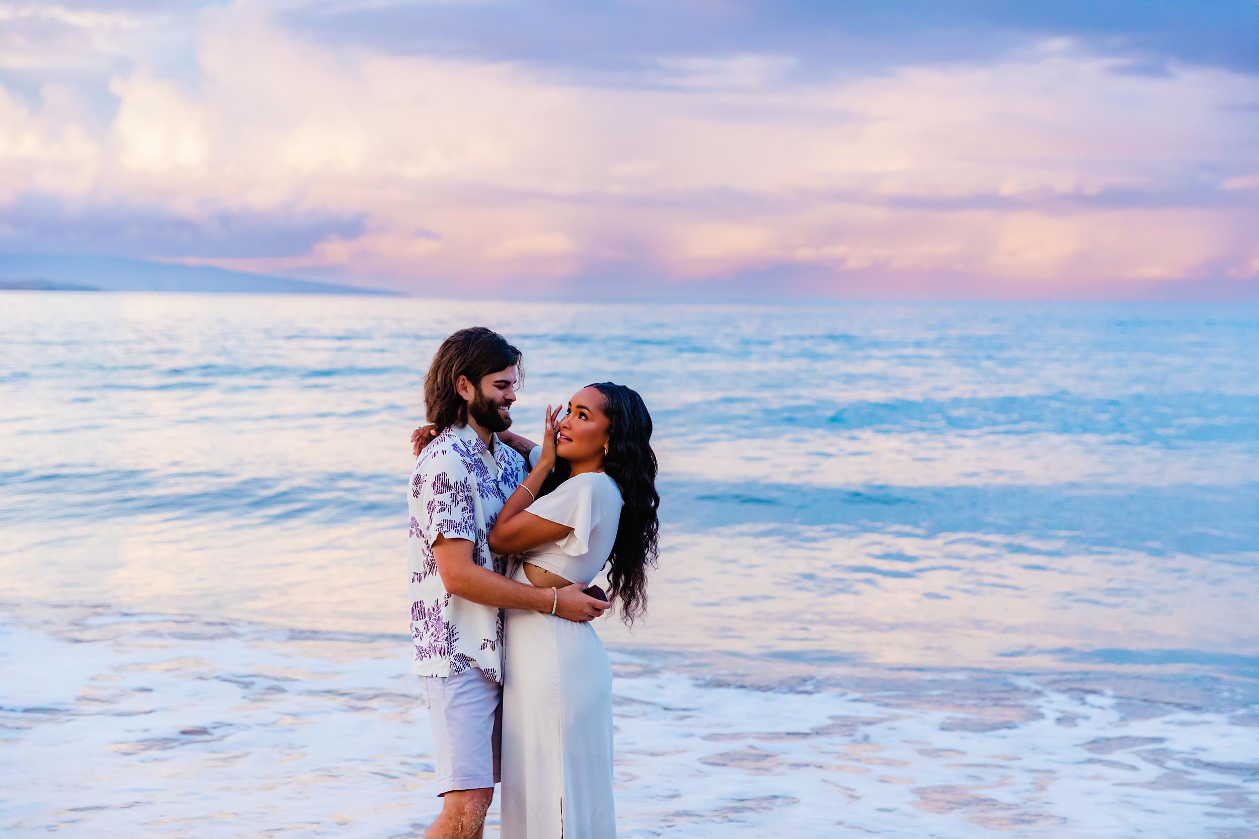 Woman with long black wavy hair and white dress wipes a tear from her eye after her boyfriend proposes to her at sunrise in Hawaii. Boyfriend has beard and patterned white shirt and smiles while he embrases her.