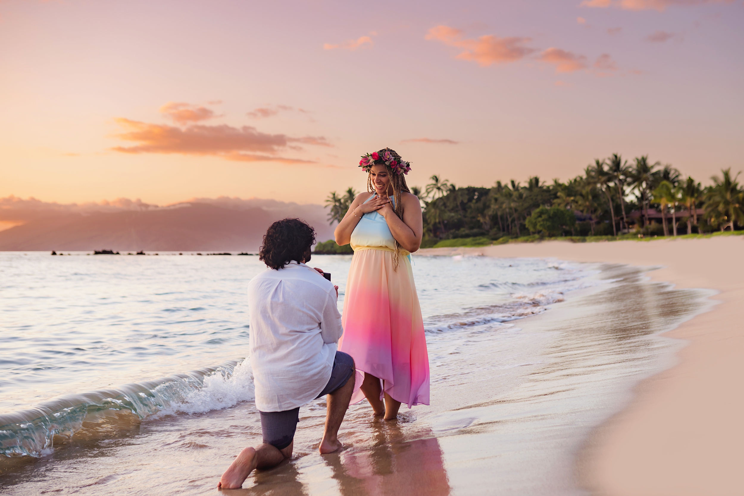 Woman wearing red lei po'o holds her chest in surprise after her boyfriend proposes to her at sunset.