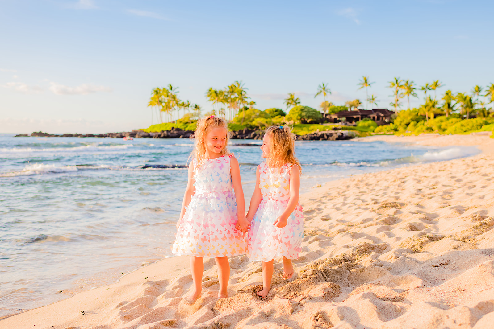 Two young girls wearing white dresses with colorful butterflies walk hand-in-hand in the sand during a Maui family photography session on the beach.
