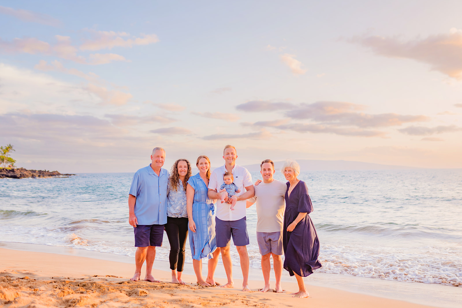 A family wearing white and blue stands together for family beach pictures on Maui, Hawaii at sunset.