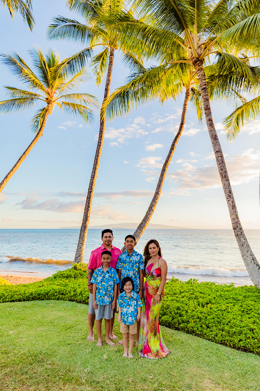 A young family wearing fun family photo beach outfits pose in front of palm trees during a photo session with Love + Water Photography on Hawaii.