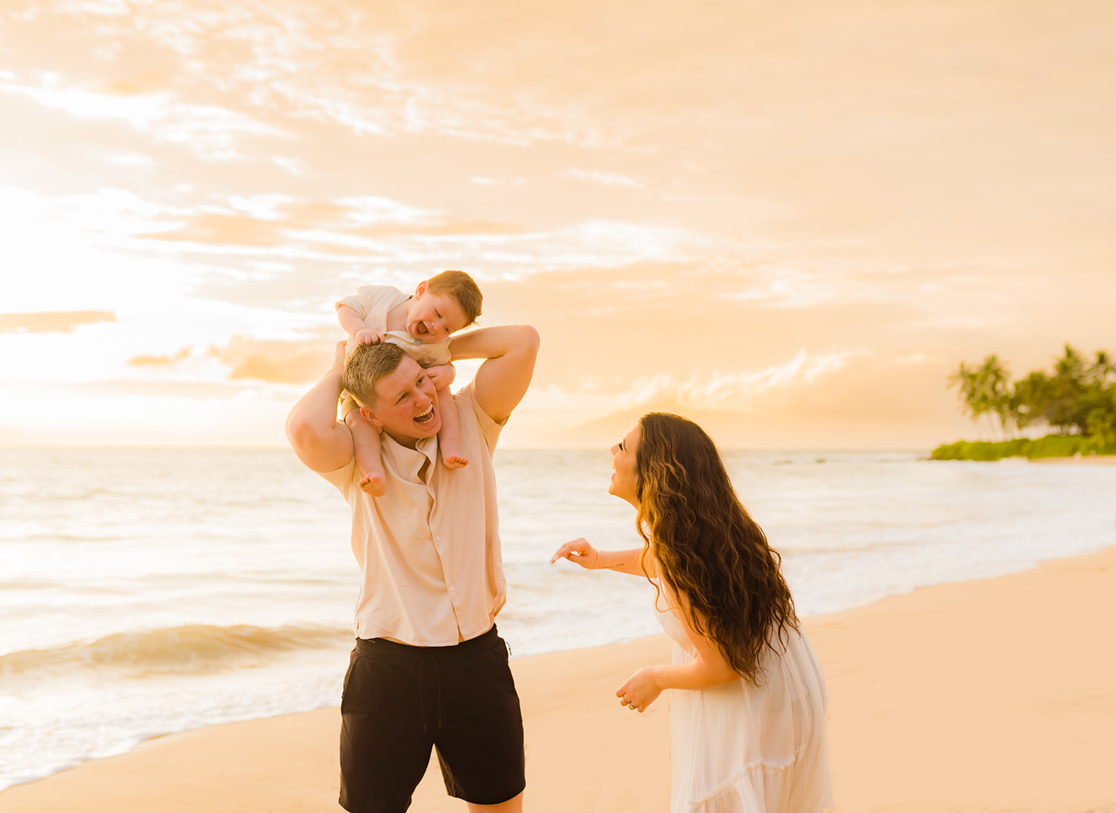 A father piggybacks his young child while the mother watches and laughs during a family beach photo session on Hawaii with Love + Water Photography.