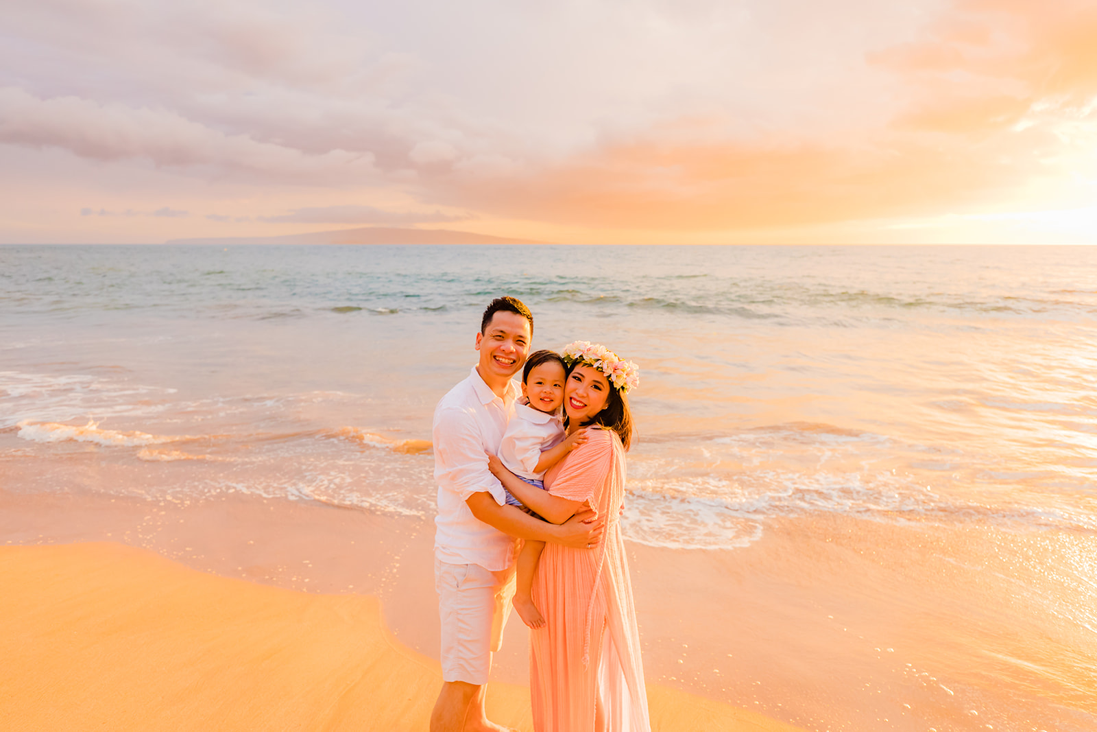 A family of three with a toddler smile at the camera during a sunset family portrait session in Wailea, Maui by Love and Water Photography.