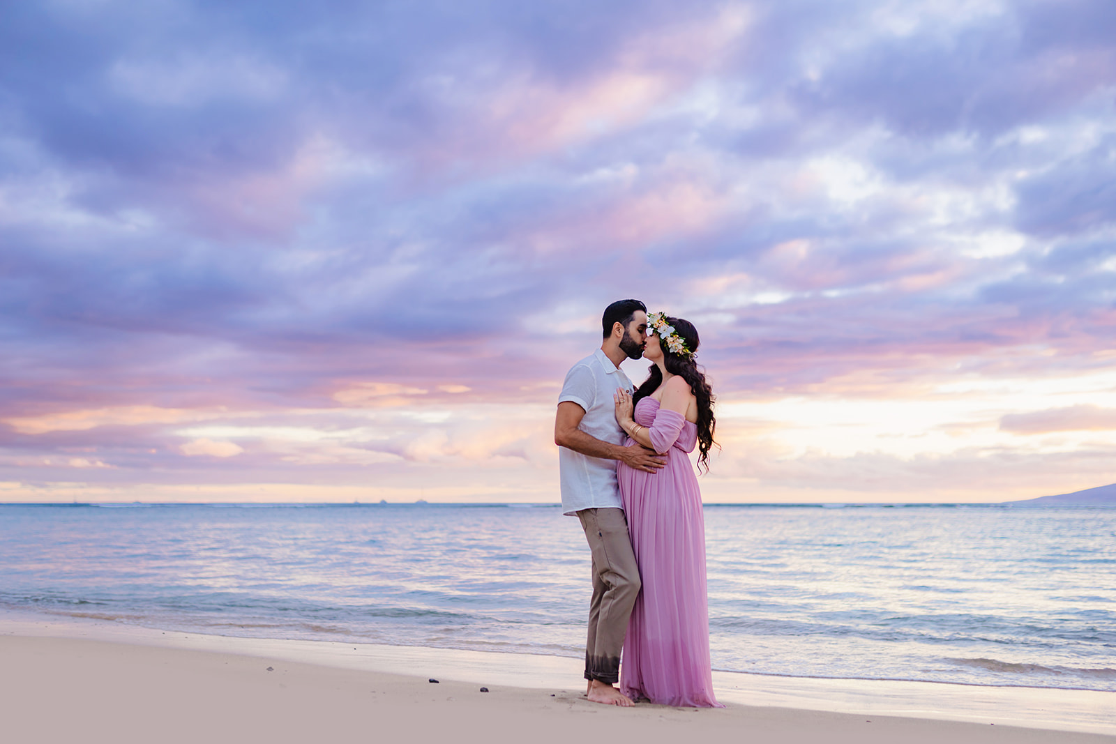 man in white shirt and beige pants kisses his pregnant wife on the beach at sunset in West Maui