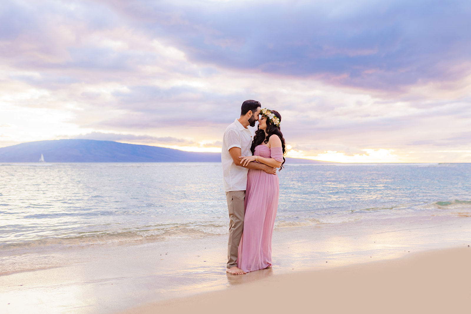 couple embraces one another under a pastel sky on the beach in West Maui during their babymoon photoshoot