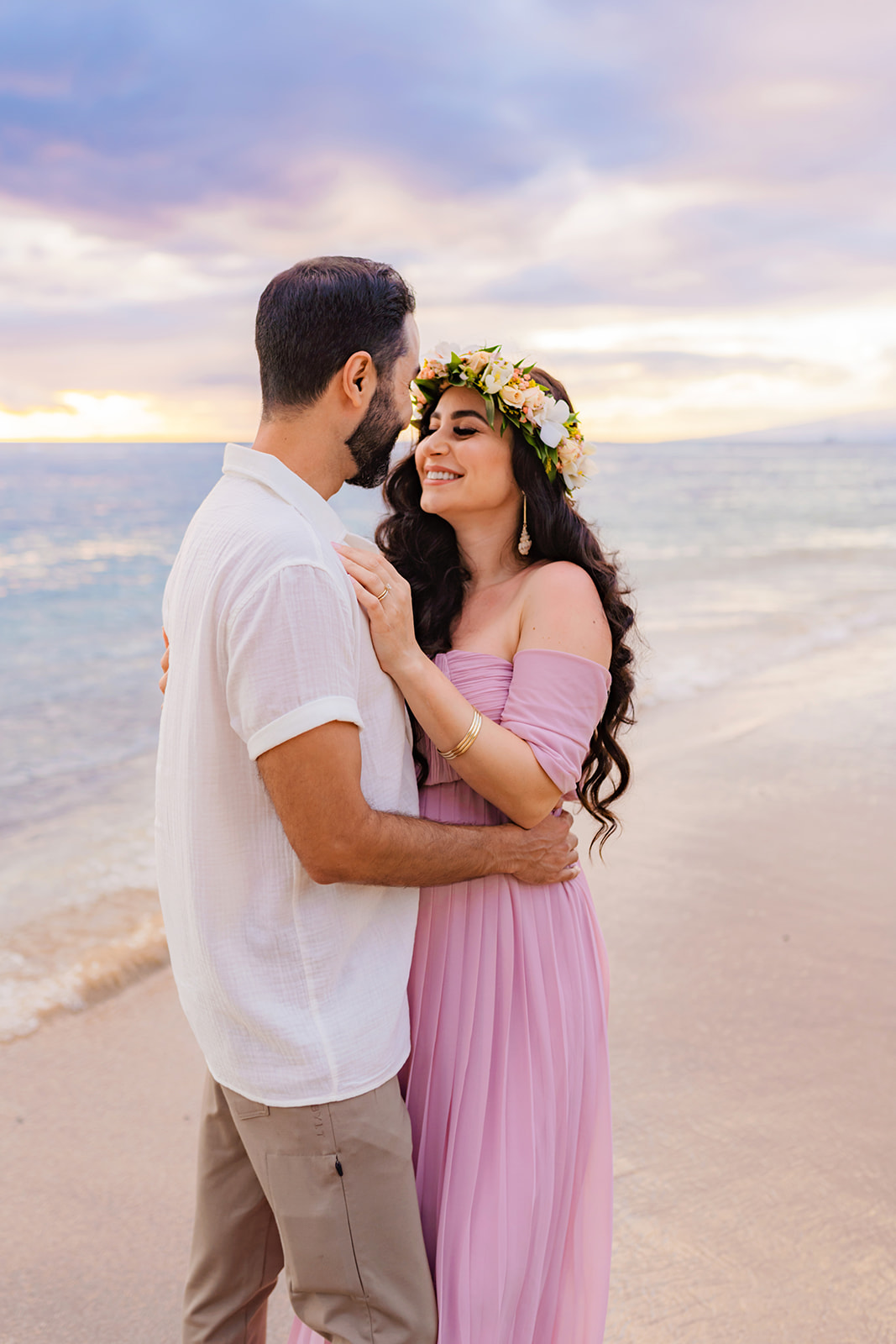 mom-to-be in a flower crown and pink dress flashes a smile at her partner at sunset on a West Maui beach