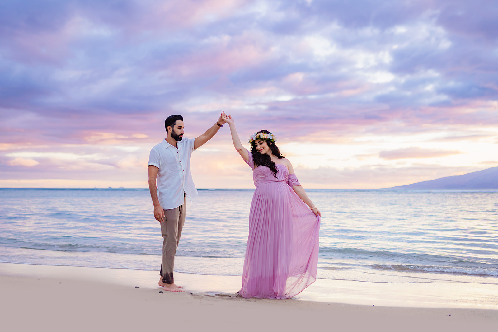 Man in white button-up shirt twirls his pregnant wife on the beach under a pink and blue pastel sunset in West Maui