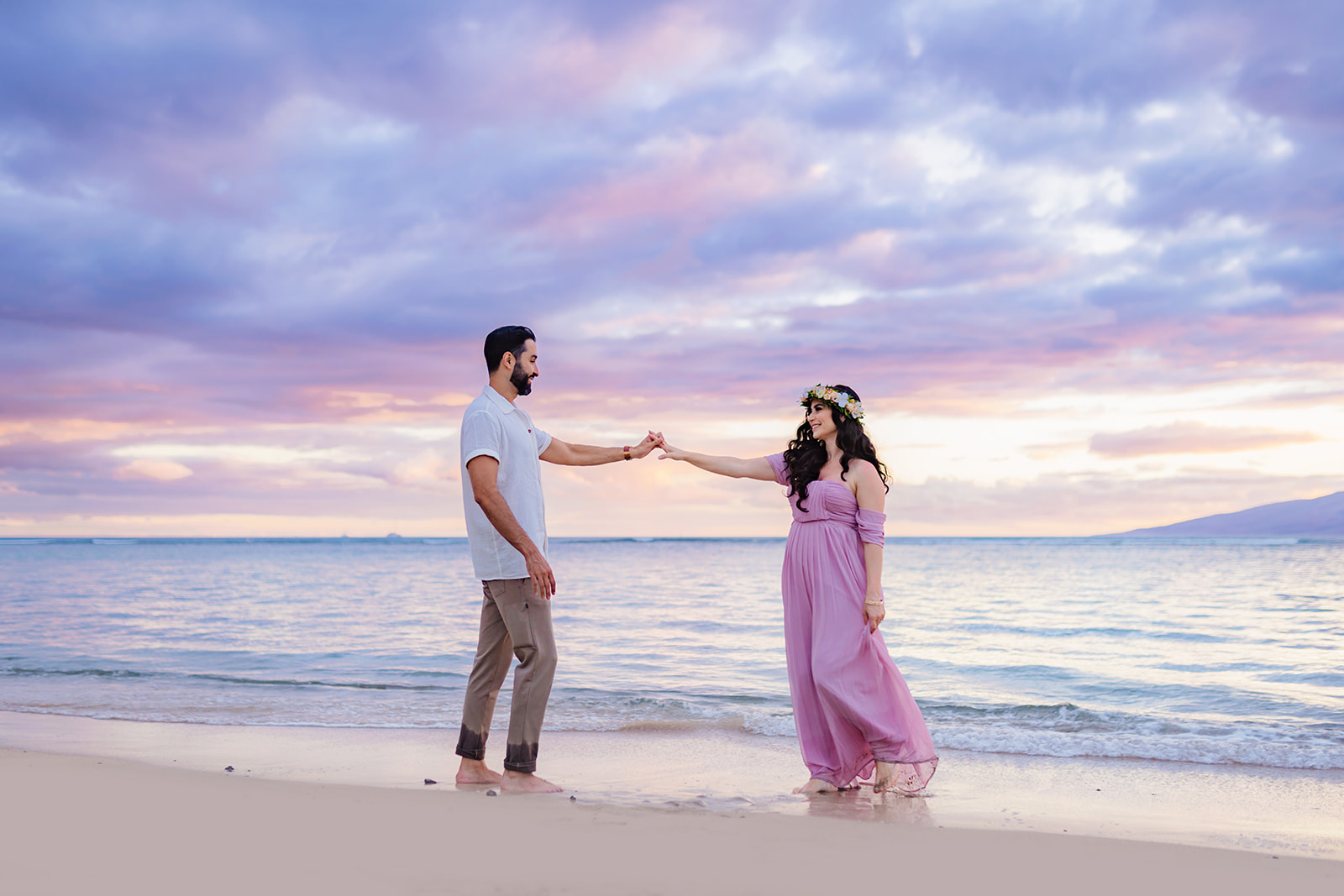 Man in white button-up shirt dances with his pregnant wife on the beach under a pink and blue pastel sunset in West Maui