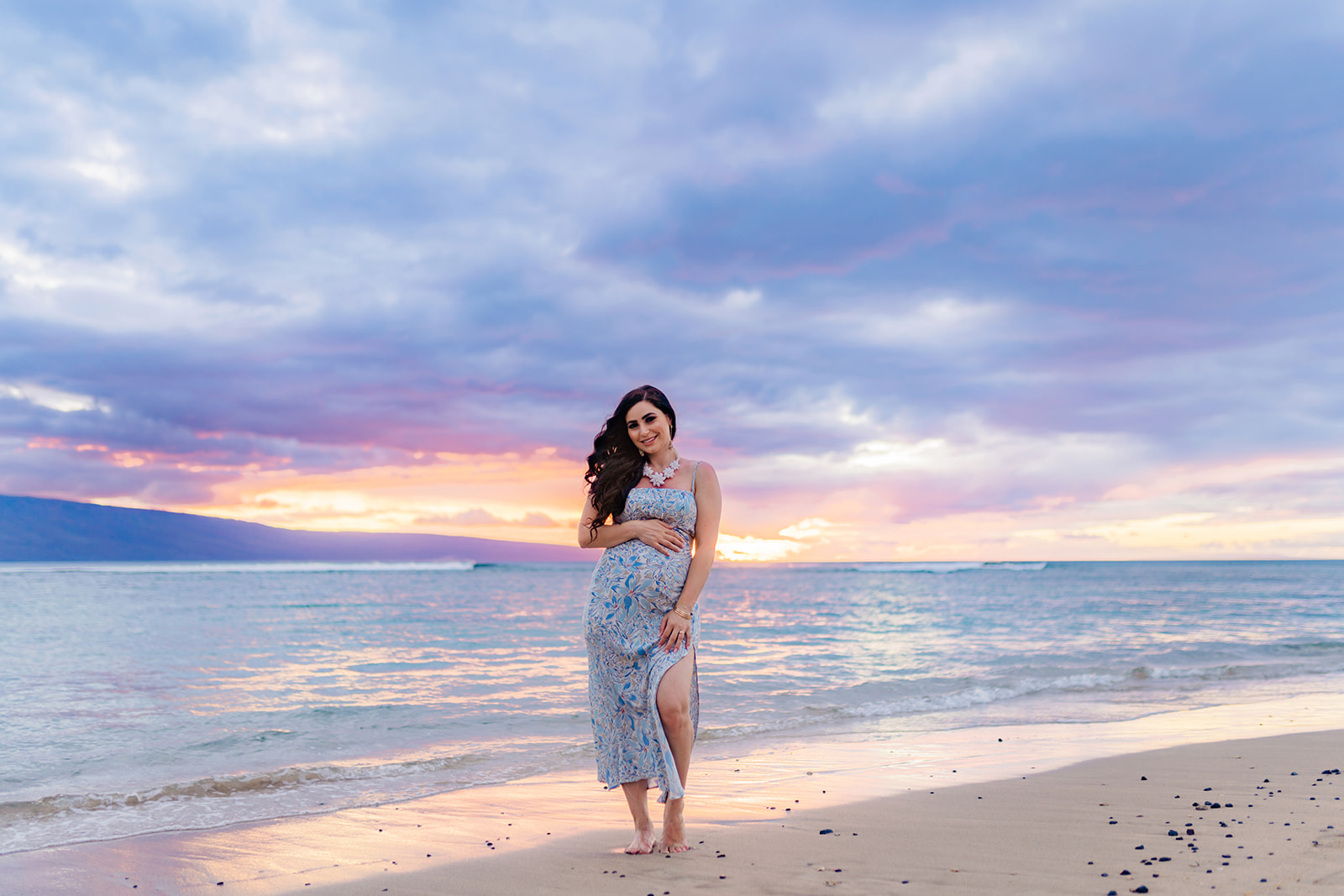 Long-haired brunette woman wearing a chunky necklace and blue and white  dress poses on the beach at sunset