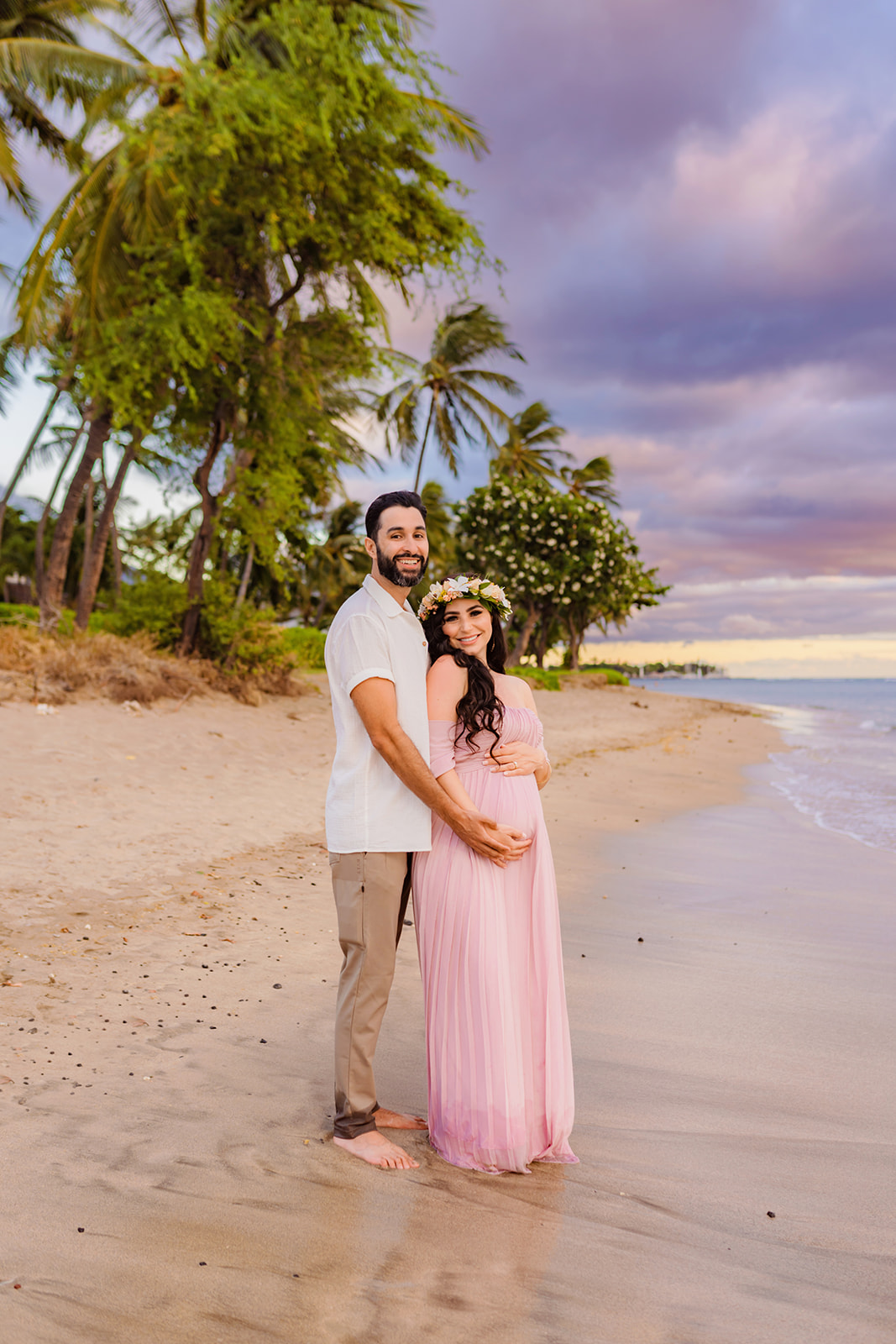 man stands on the beach barefoot in a white shirt and beige pants holding the baby bump of his partner standing in a pink dress with him