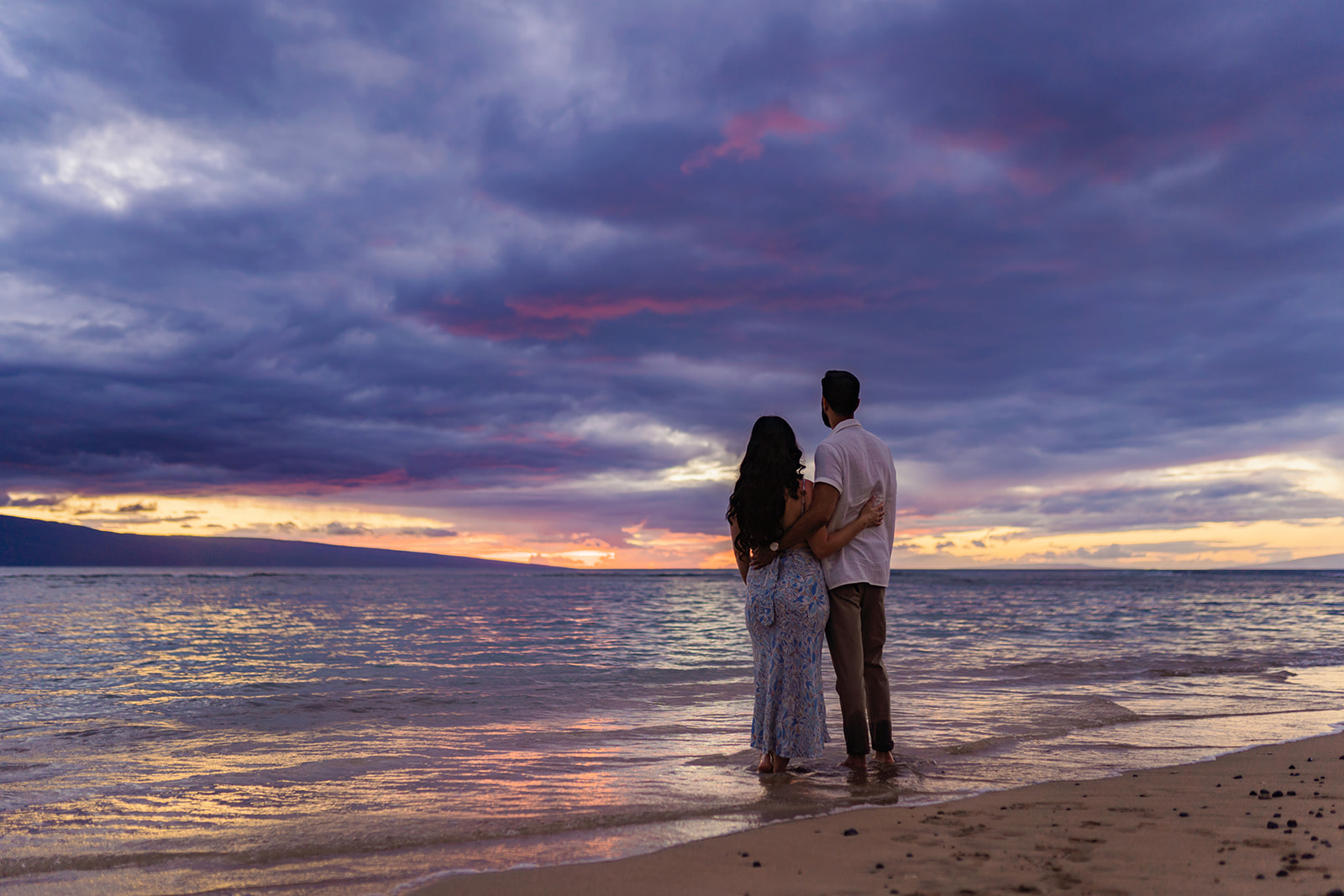 Man and woman stand faced away from the camera looking out towards the West Maui sky at sunset 