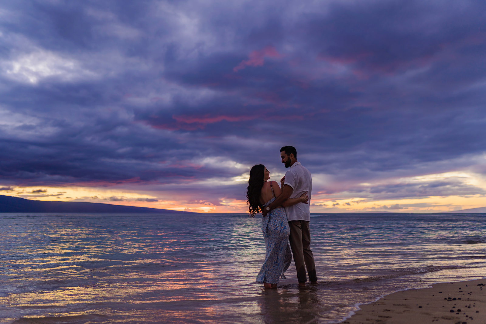 Couple laughs together during their maternity shoot with their toes in the sand as the sun sets in West Maui