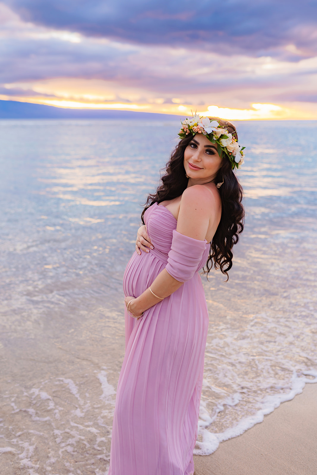 woman in off-shoulder pink dress caresses her baby bump while smiling at the camera of her maternity photographer in west Maui