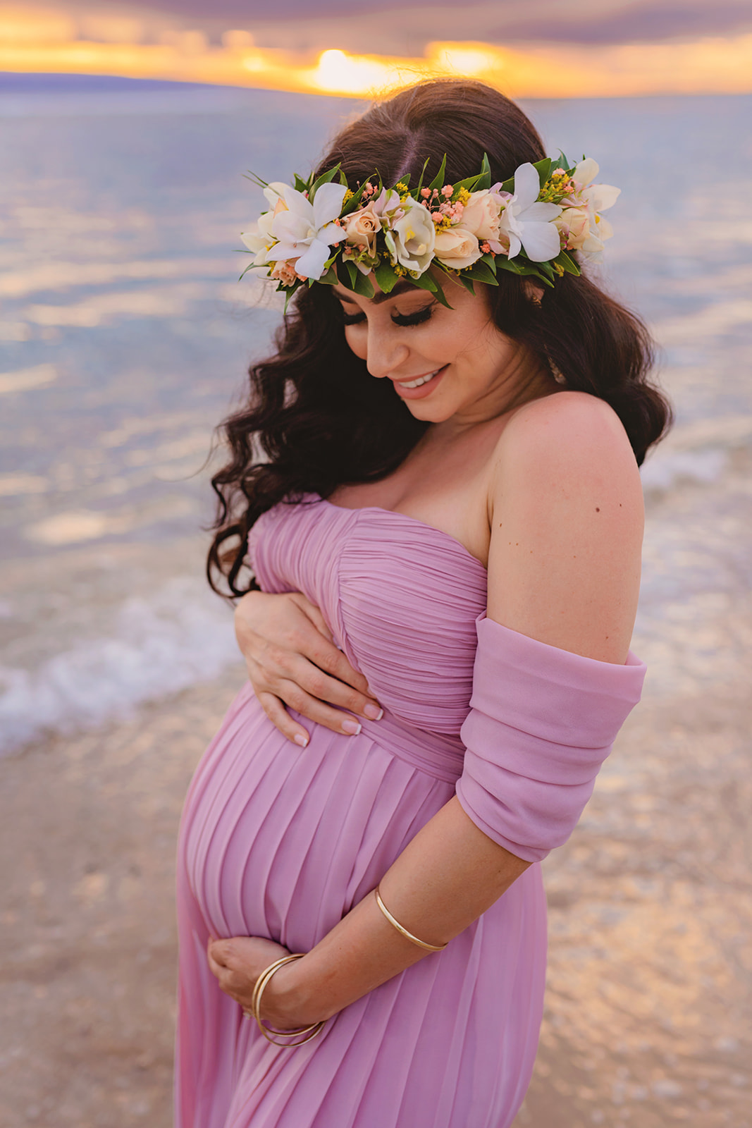 long-haired brunette woman in a flower crown holds her baby bump and smiles on the beach at sunser