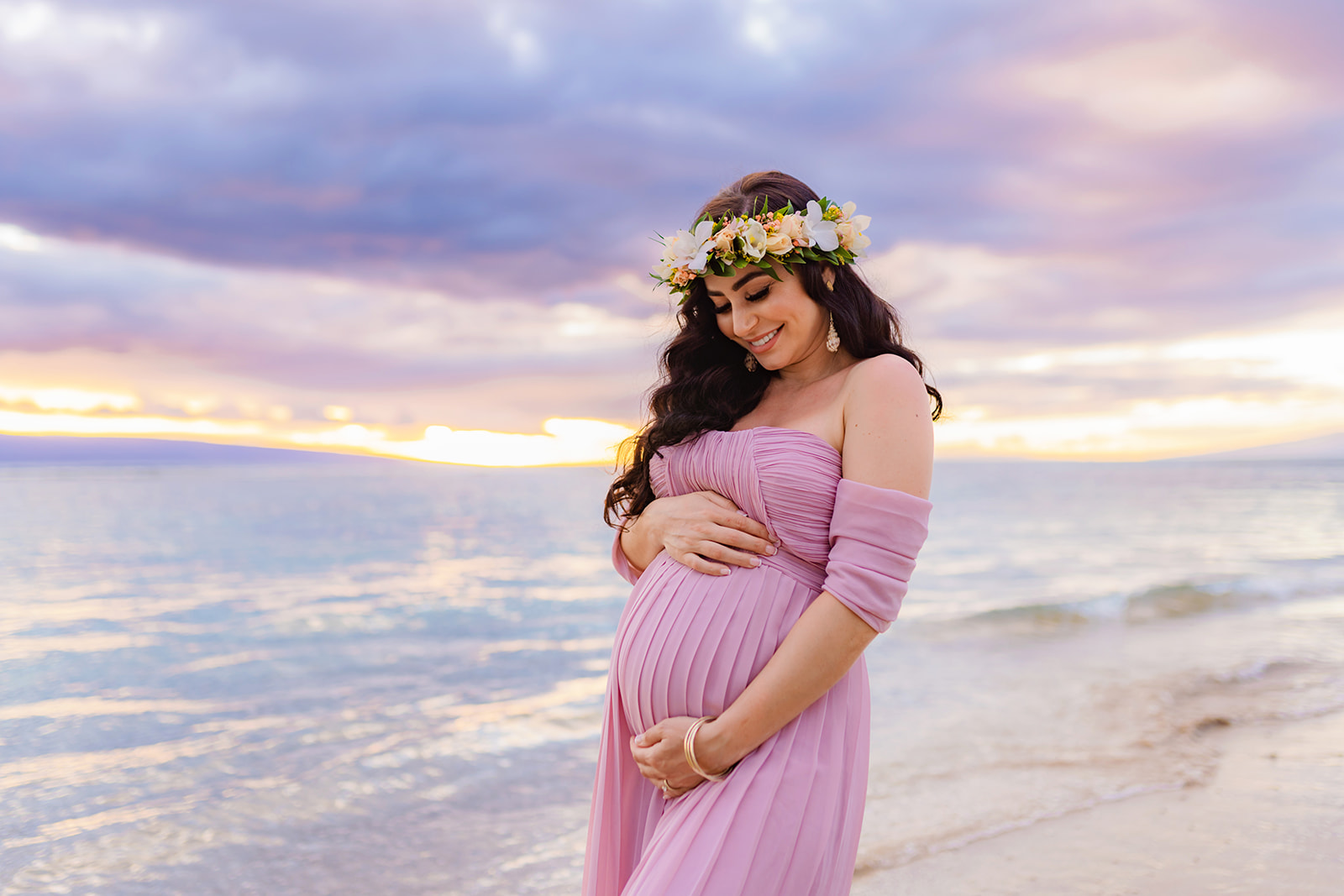 woman in a flower crown smiles down at her baby bump as she stands in a flowy pink maxi dress on a West Maui beach