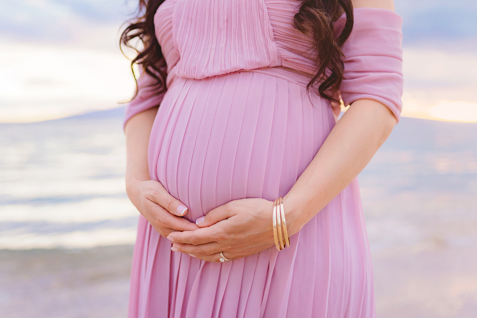detail shot of a woman holding her baby bump from underneath in a pink dress and wearing gold bangles around her wrist