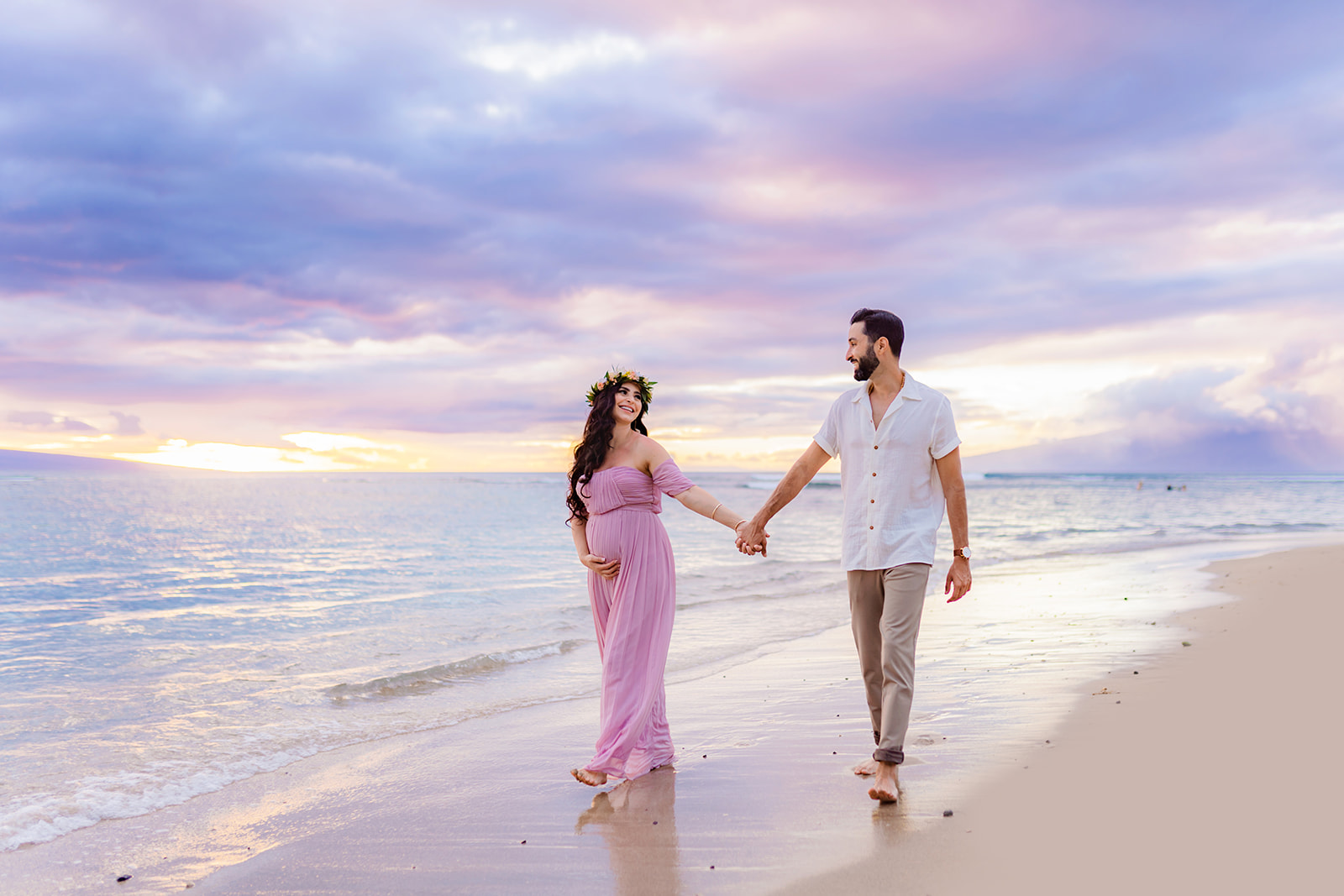 pregnant woman strolls on the beach with her significant other hand in hand smiling under a pastel sky