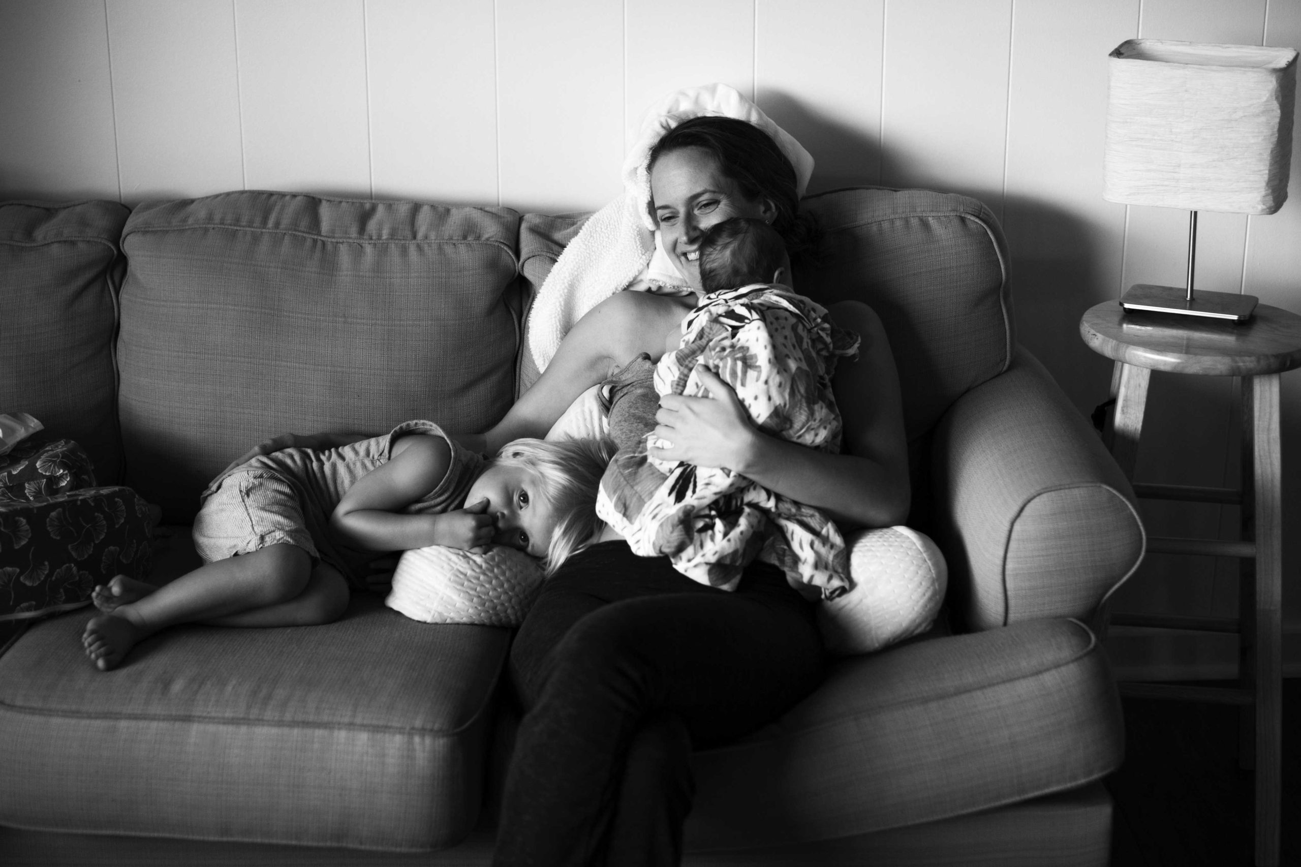 Black and white image of new mother holding her newborn to her chest on the couch with toddler boy snuggled next to her.