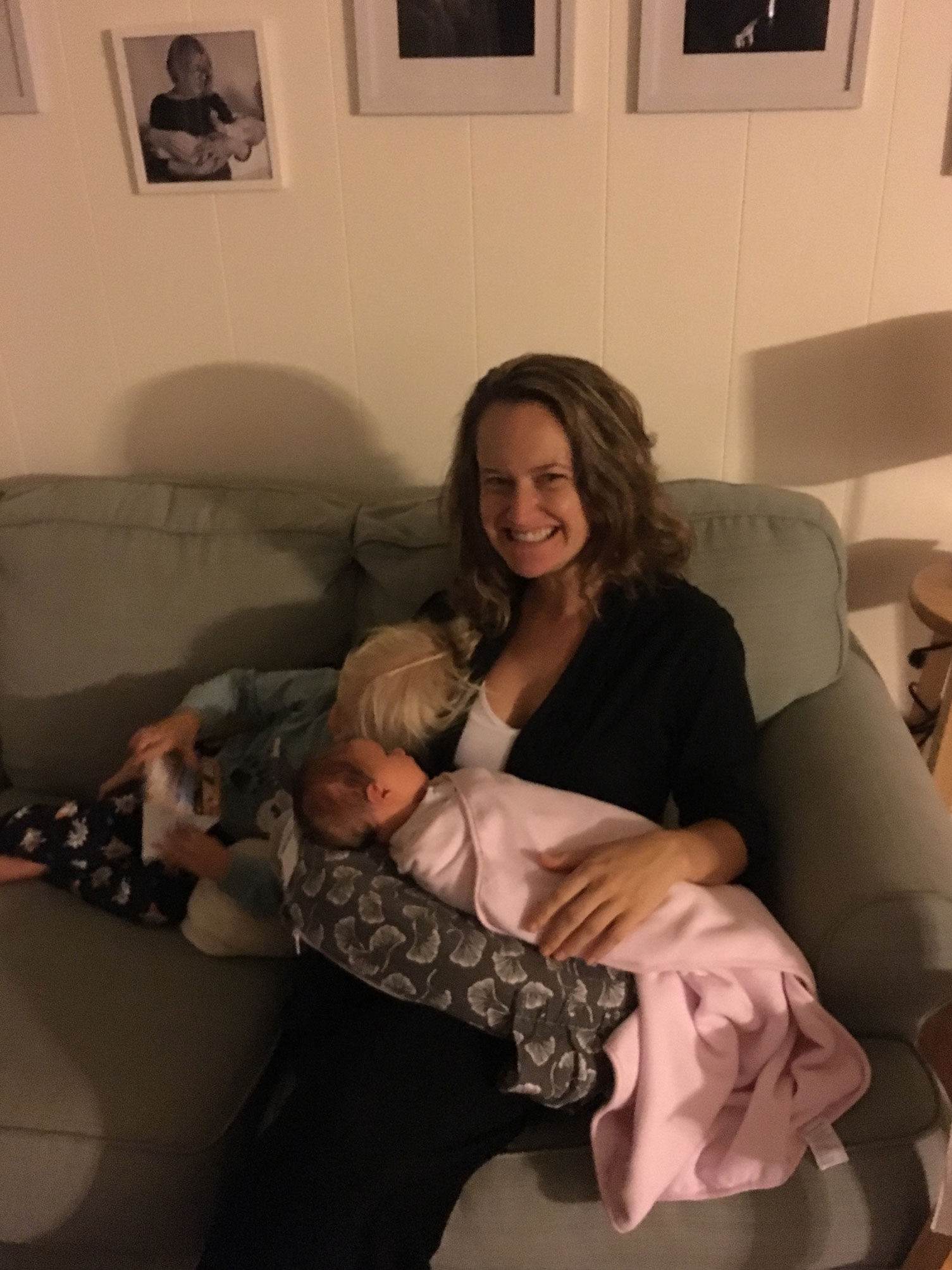 New mother holds newborn on the couch during postpartum recovery period while cuddling her toddler.